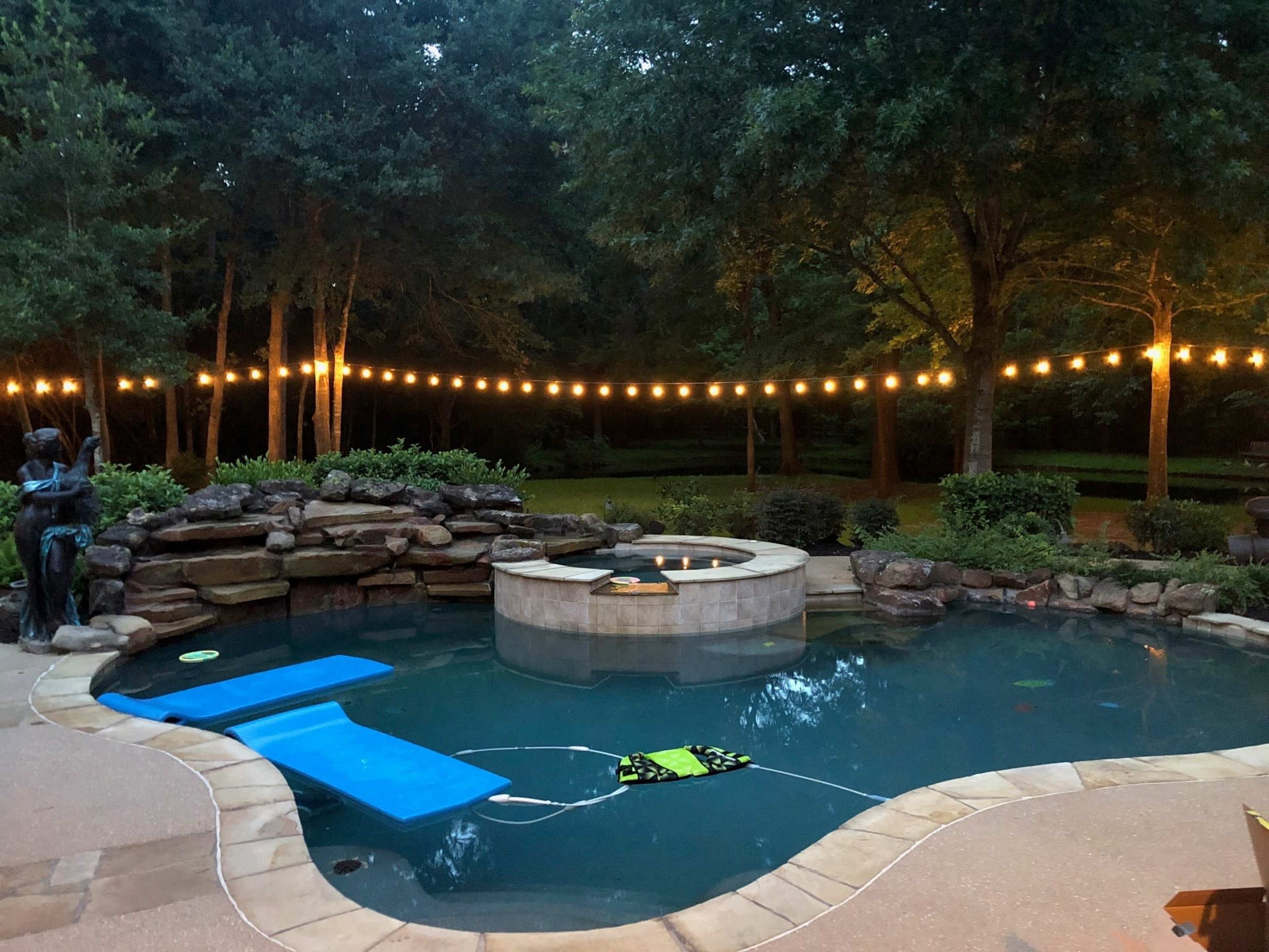 A backyard pool with a raised hot tub, surrounded by a stone wall and illuminated by string lights overhead.