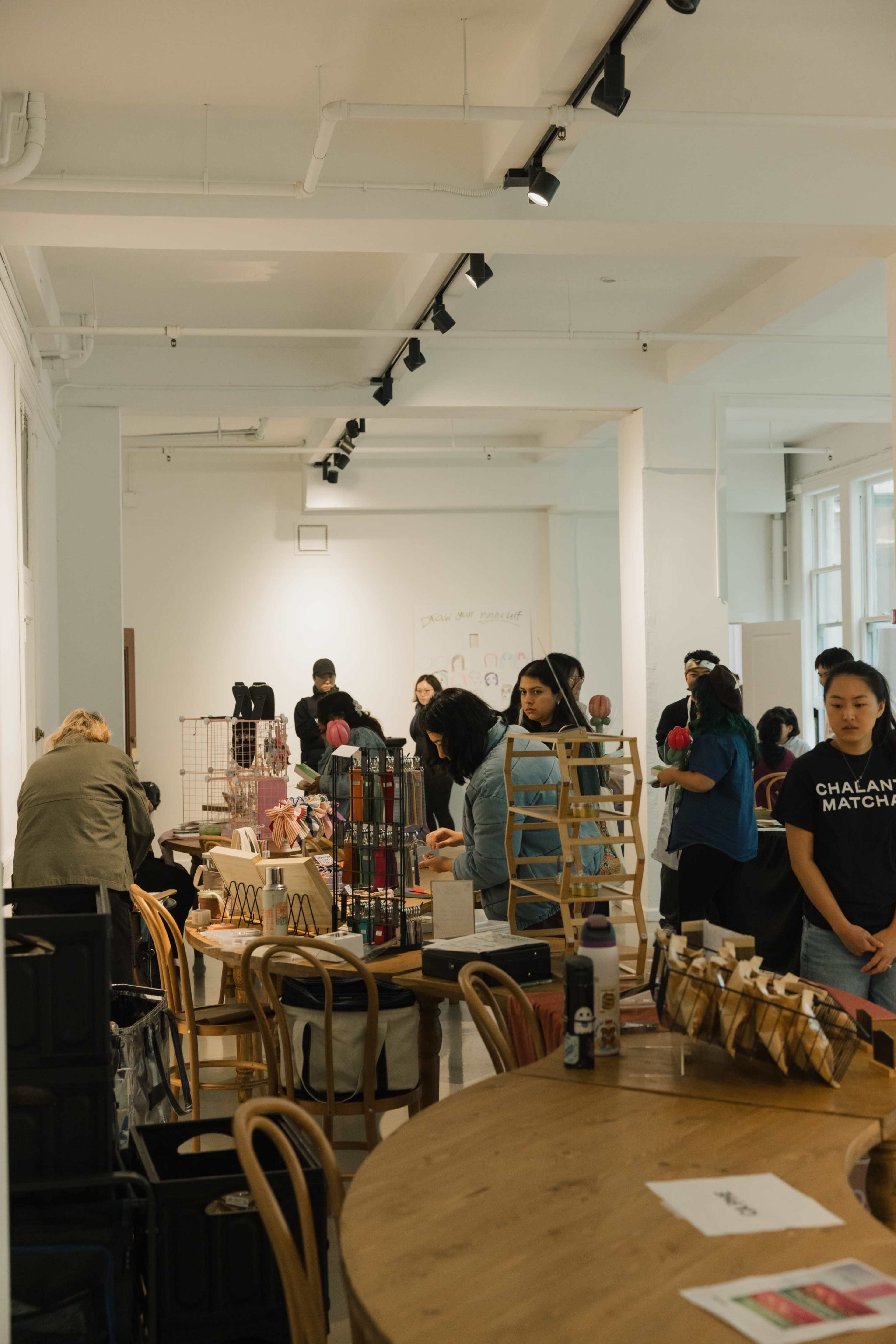The image shows a crowded indoor market event with various vendor displays, including tables filled with merchandise, while people browse and interact in the space.