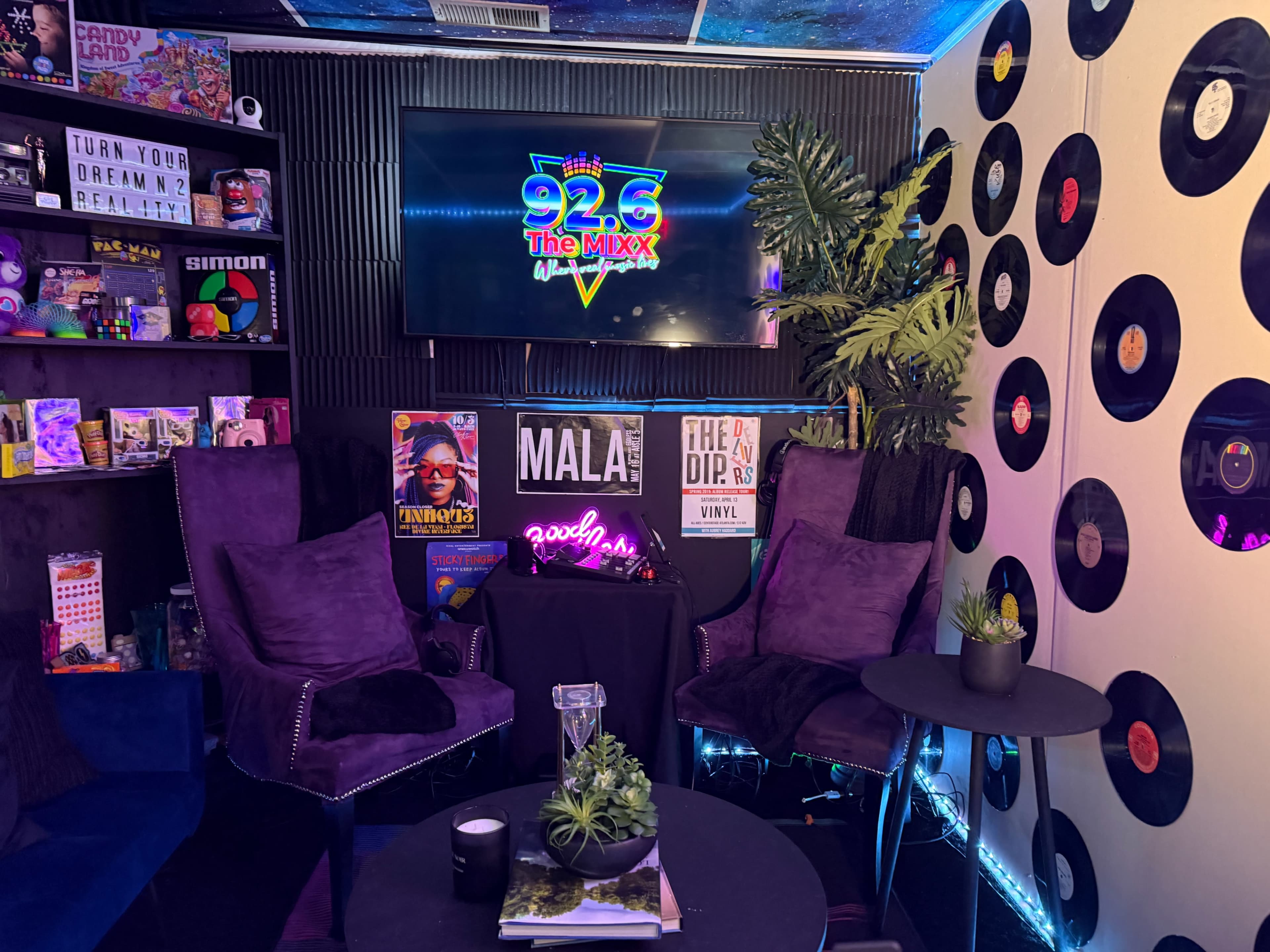 The image shows a cozy media studio with two purple chairs, a television displaying "92.6 The Mixx," and decorated walls featuring vinyl records and various colorful posters.