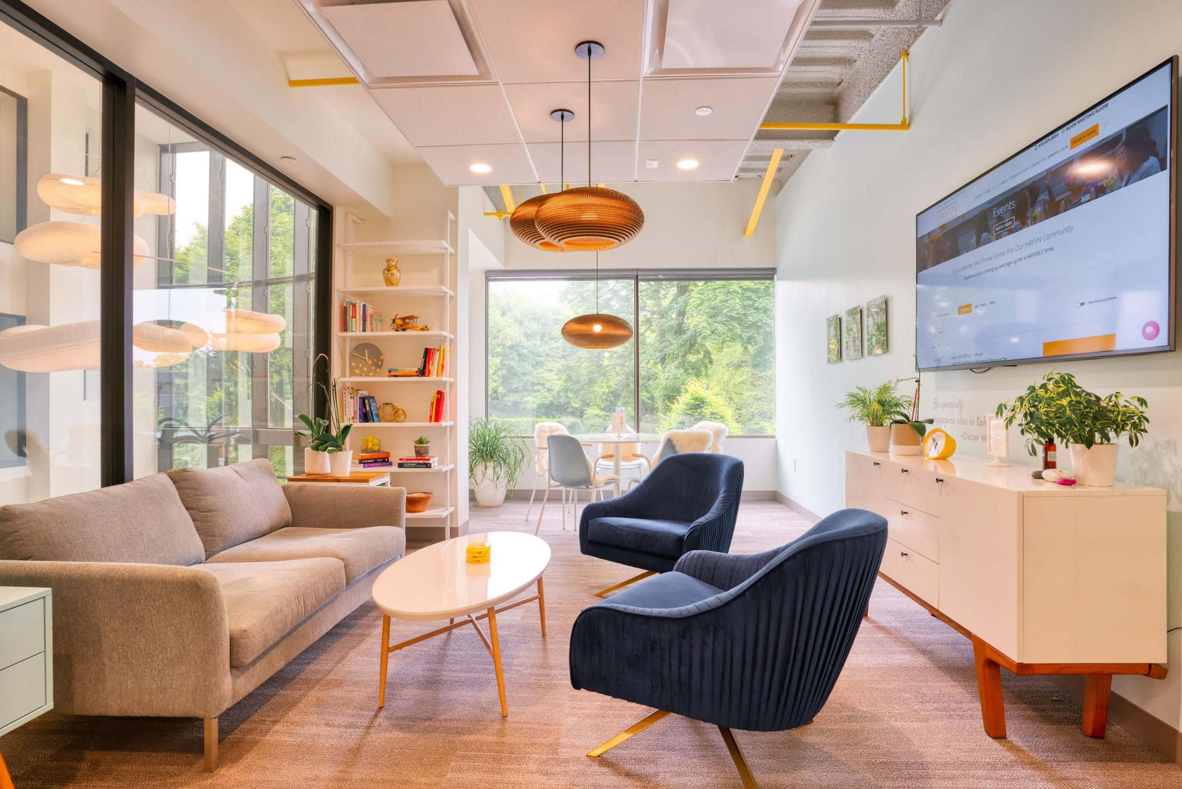 A modern office lounge area featuring a gray sofa, two blue armchairs, a round coffee table, and a large window with a view of greenery.