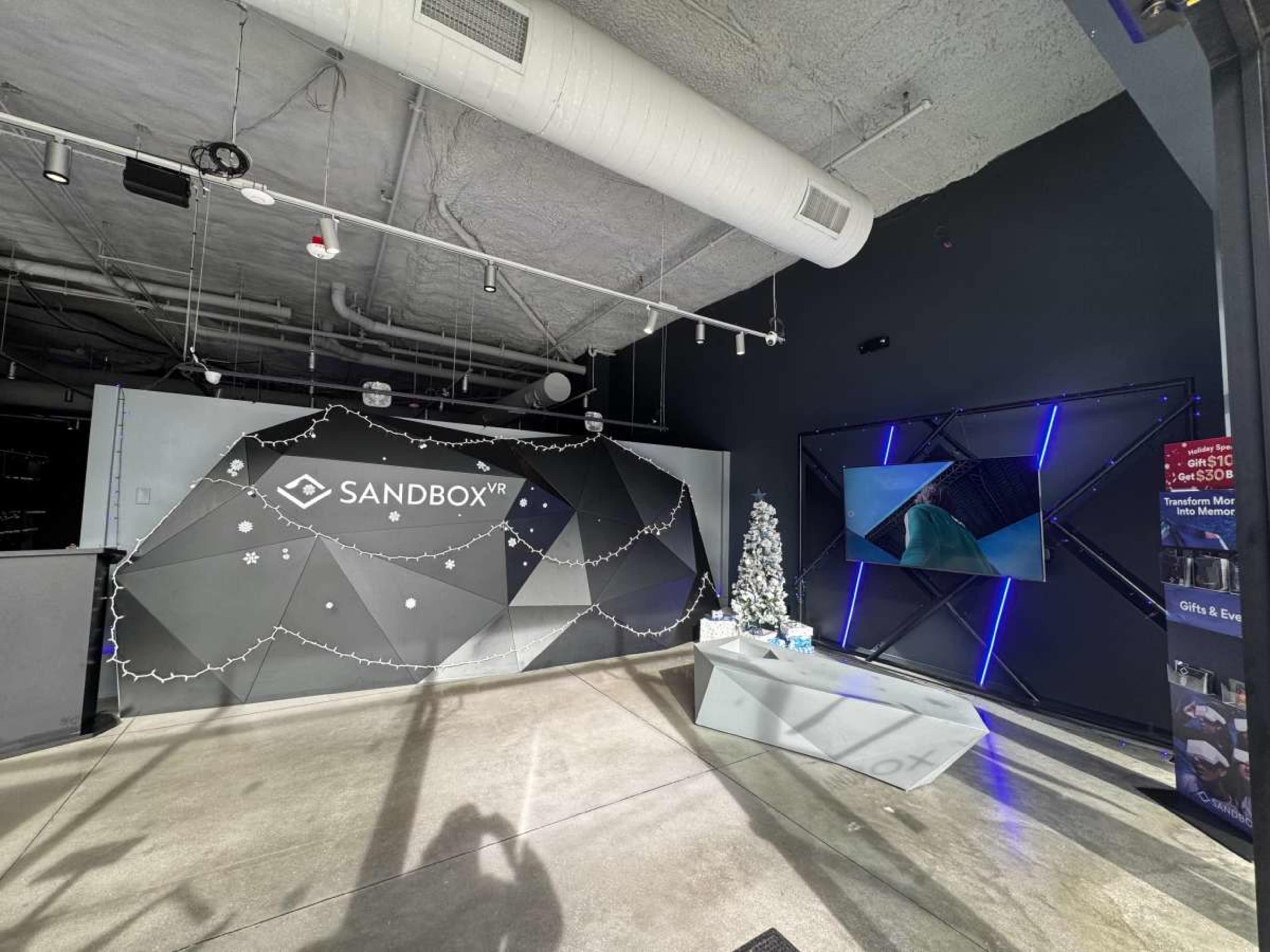 The image depicts a modern interior space with a prominent "SANDBOX VR" logo on a geometric wall, a decorated Christmas tree, and a digital screen displaying visuals on another wall.