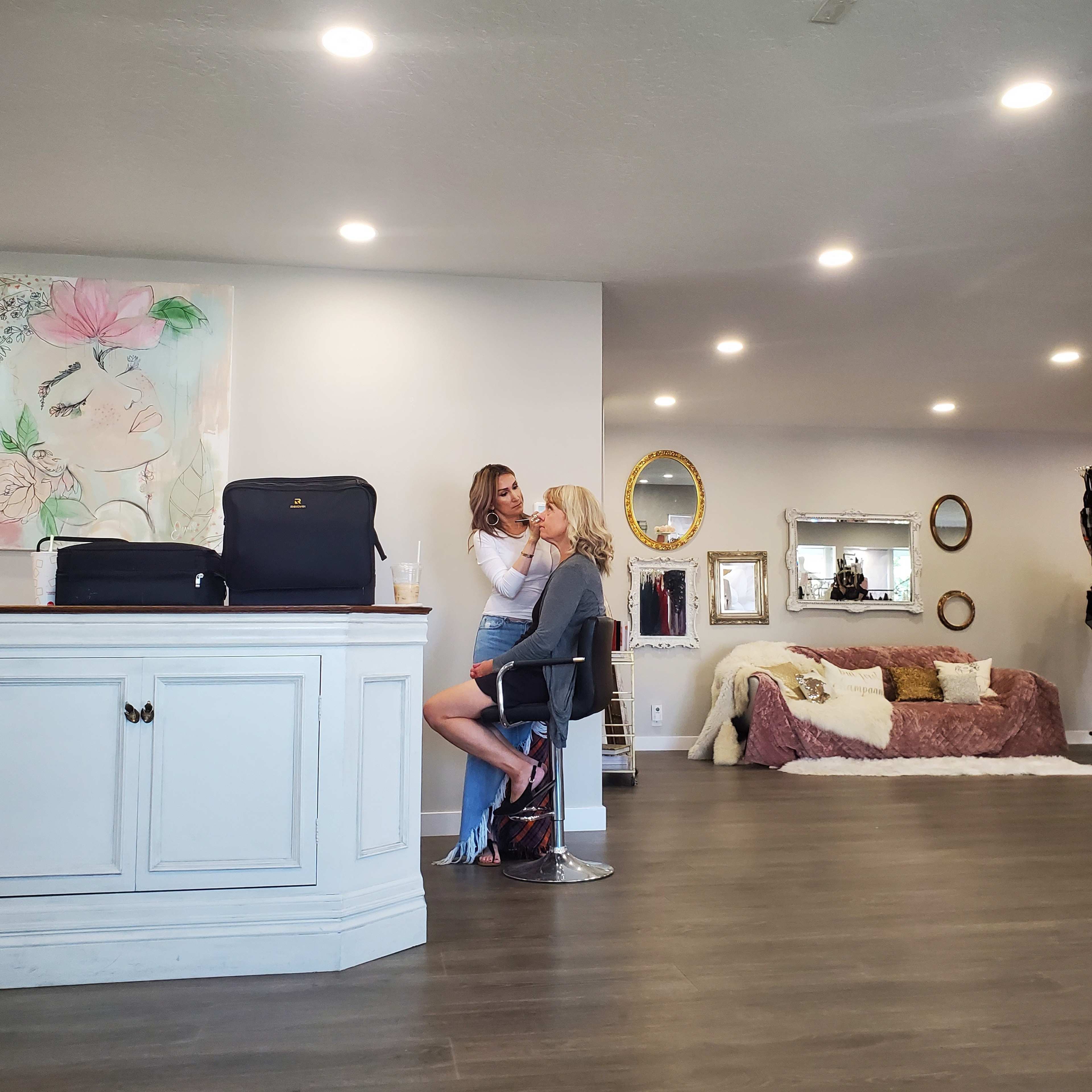 Two women are seated at a counter in a well-lit room adorned with art and mirrors, with one woman applying makeup to the other.