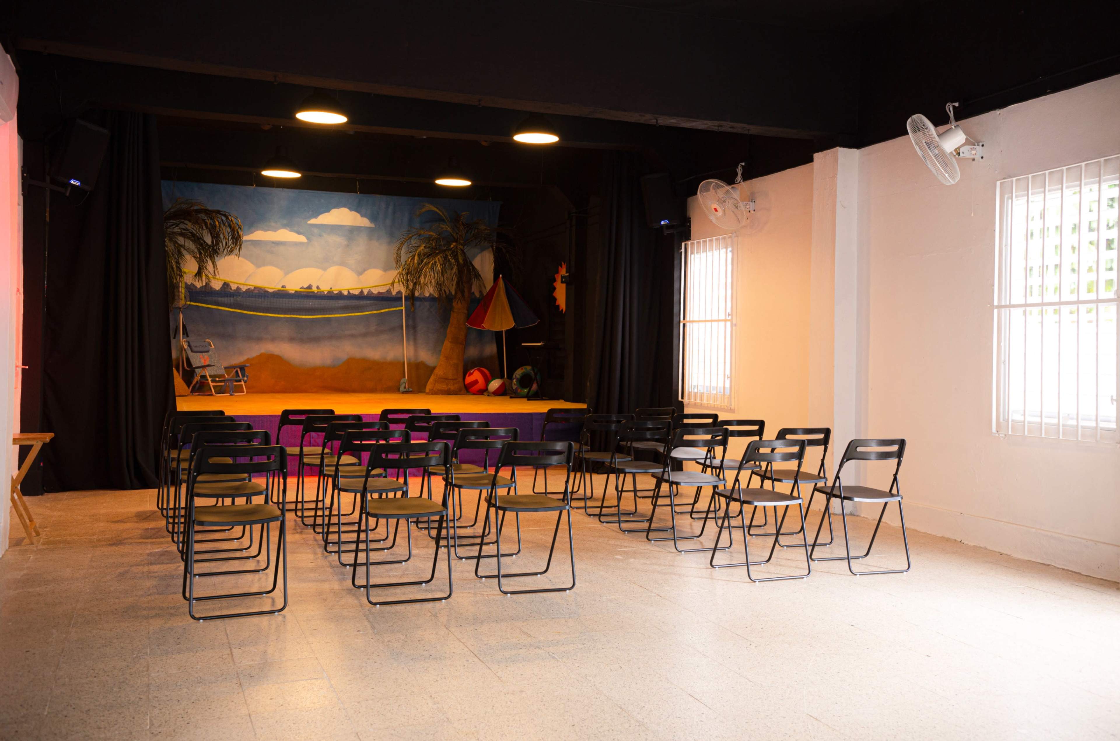 The image shows a small theater space with rows of black chairs facing a colorful stage backdrop depicting a beach scene.