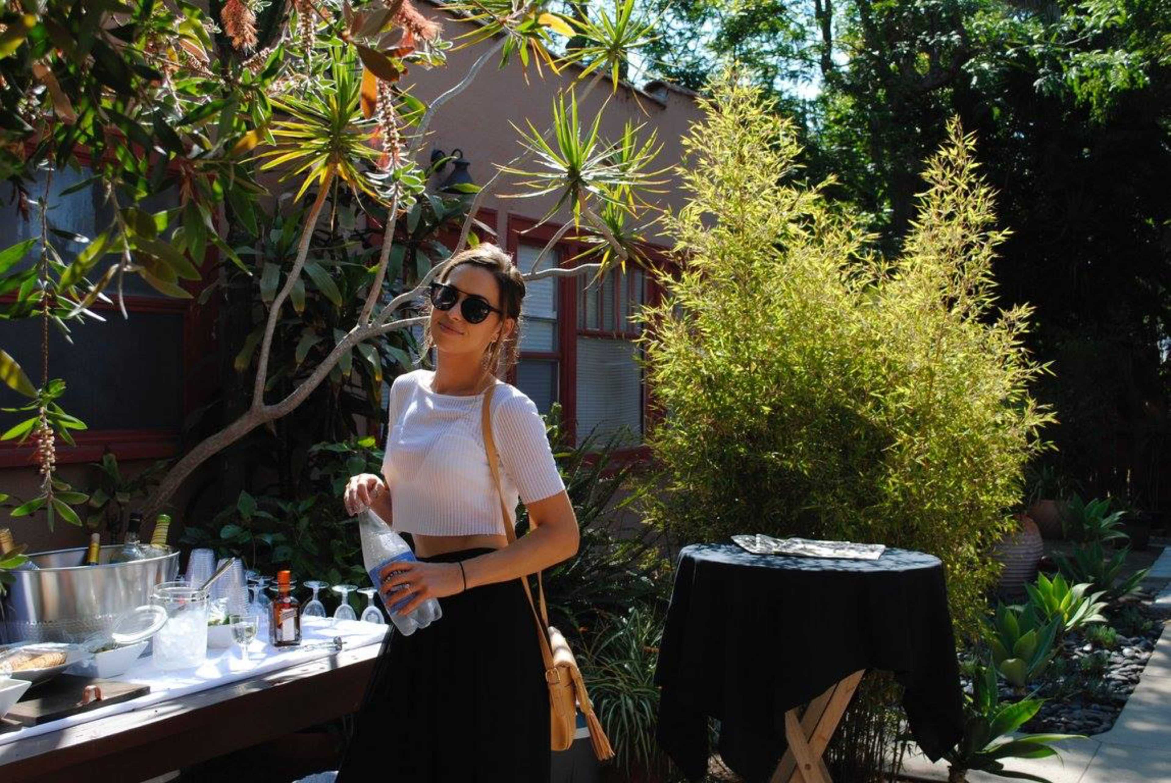 A woman stands near a table in a garden setting with tropical plants and a bamboo backdrop.