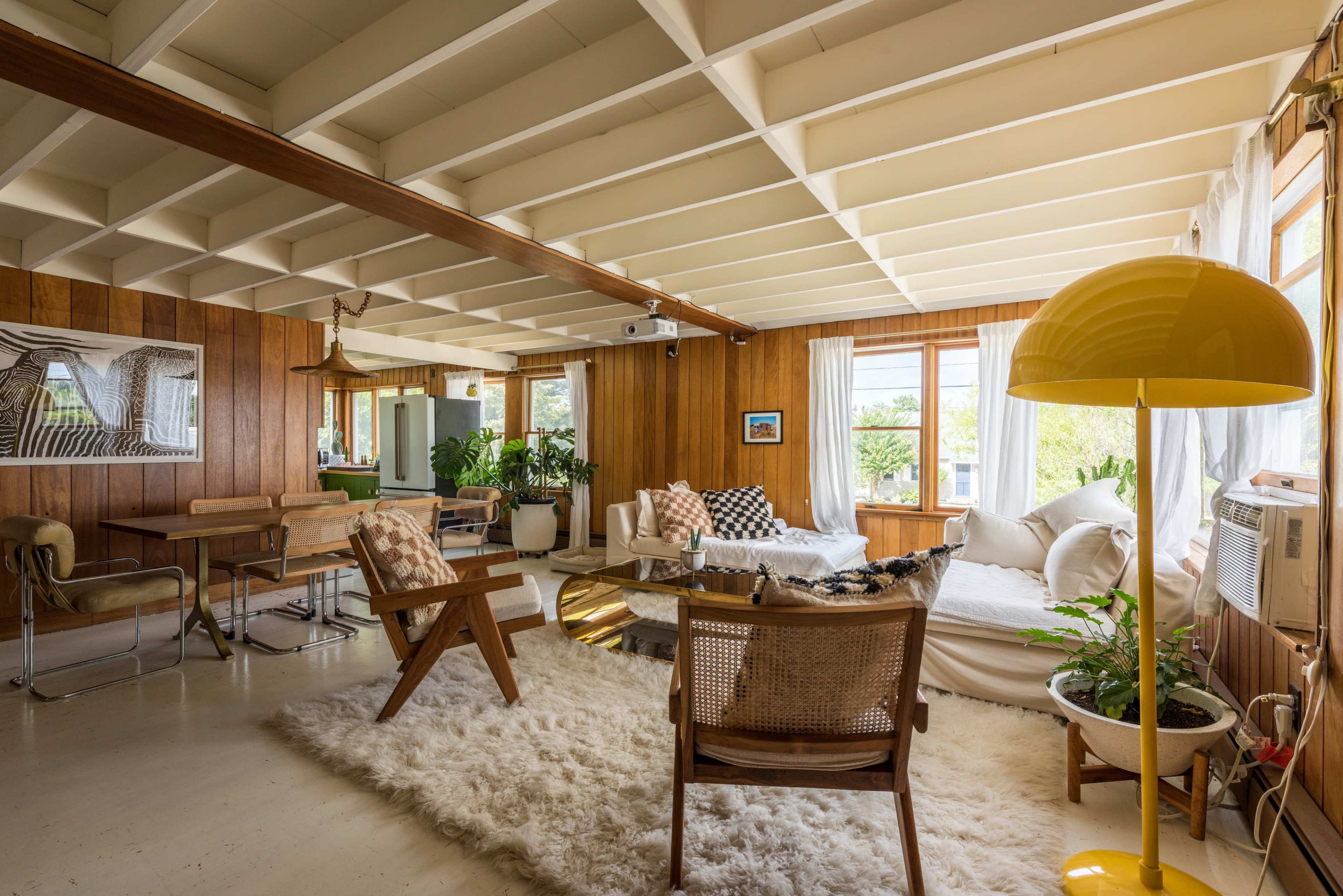 A spacious living room with wooden walls, a white rug, comfortable seating, and several indoor plants.