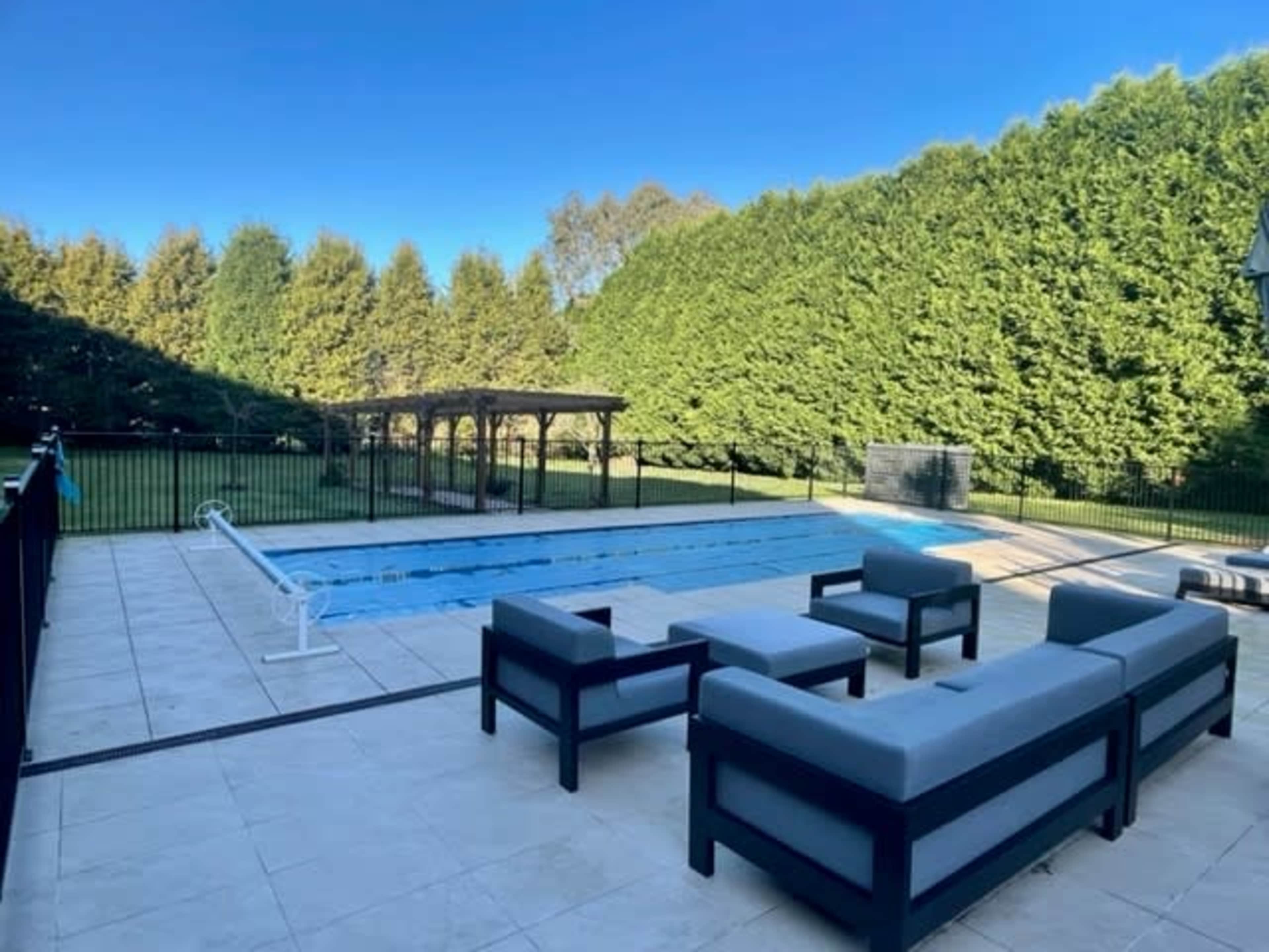 A rectangular pool with a surrounding patio and seating area is set against a backdrop of tall green hedges.
