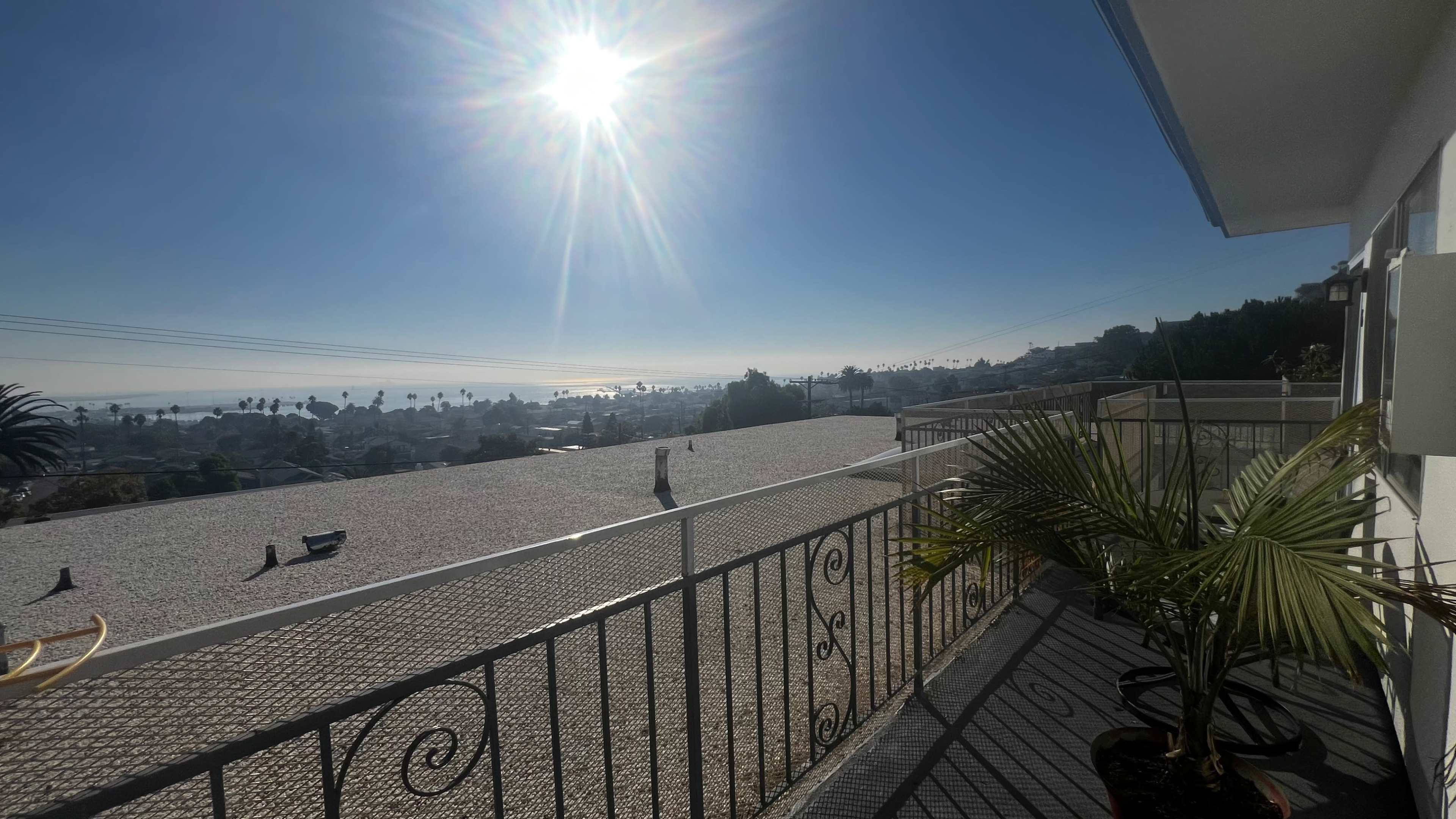 The image shows a balcony with a railing overlooking a coastal view, featuring palm trees and distant water under a bright sun.