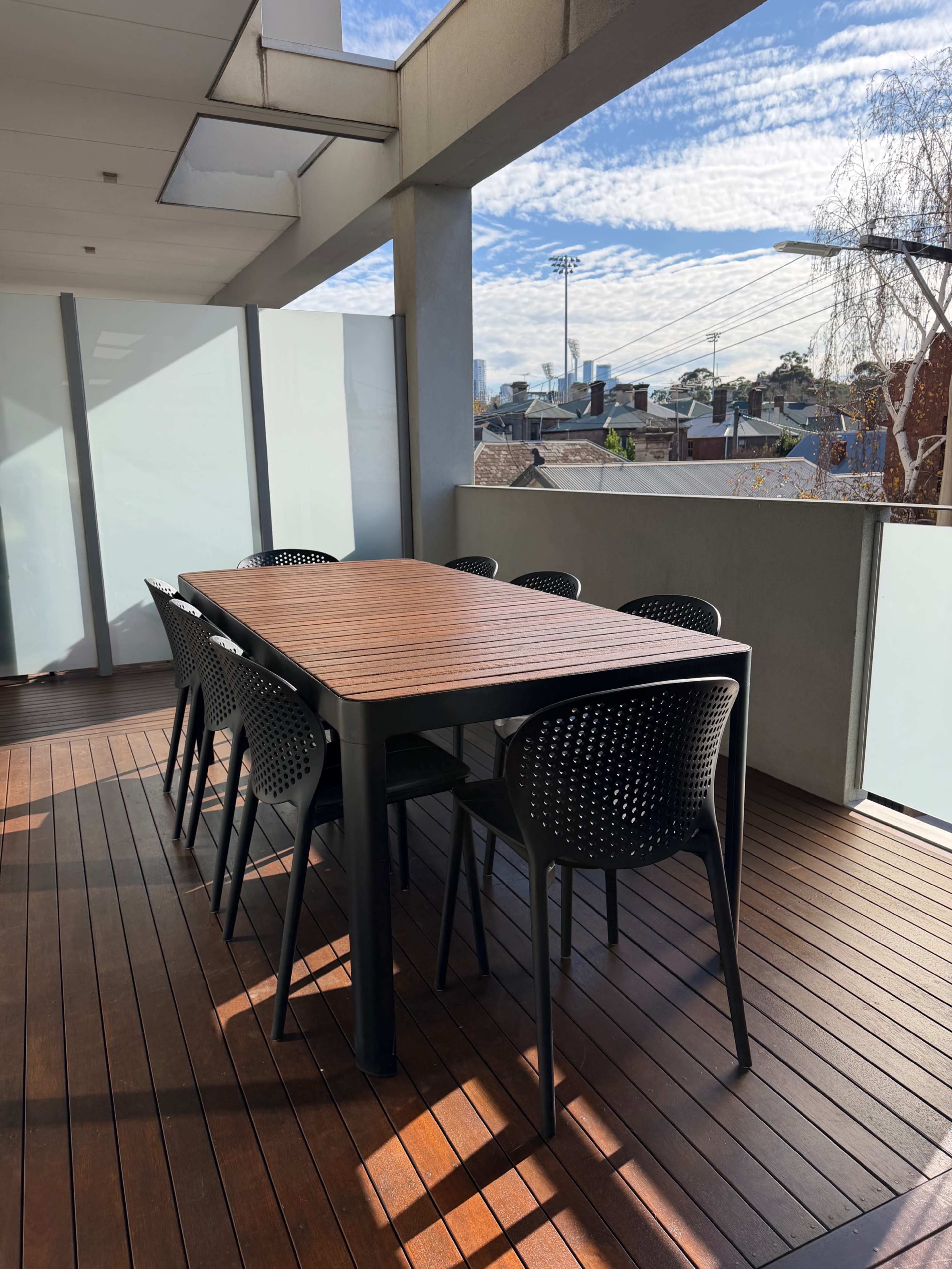A large wooden dining table surrounded by black plastic chairs is set on a balcony overlooking a neighborhood with a blue sky and wispy clouds.