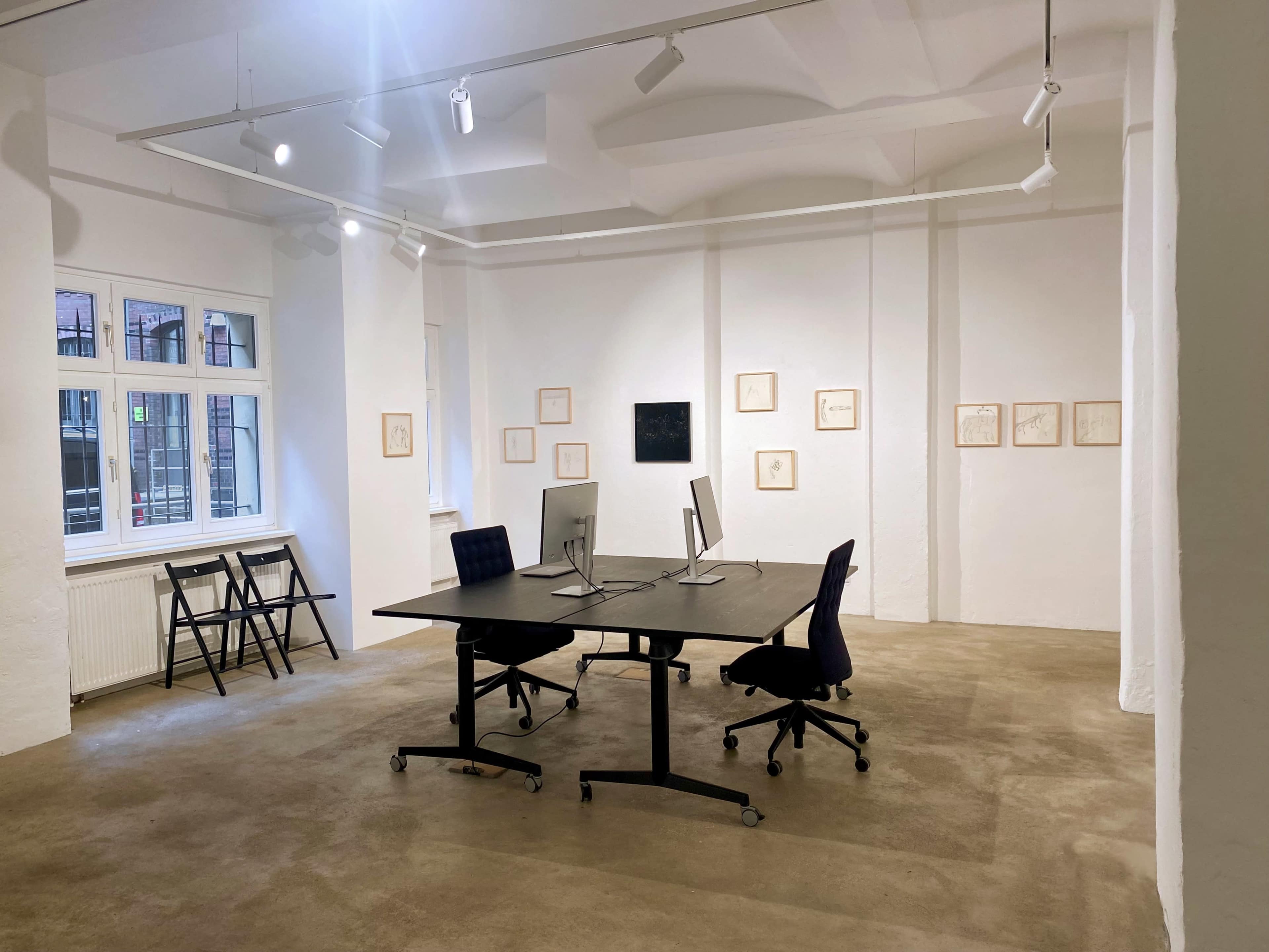 A minimalist office space featuring a large table with two monitors, surrounded by empty chairs, and artwork hanging on the white walls.