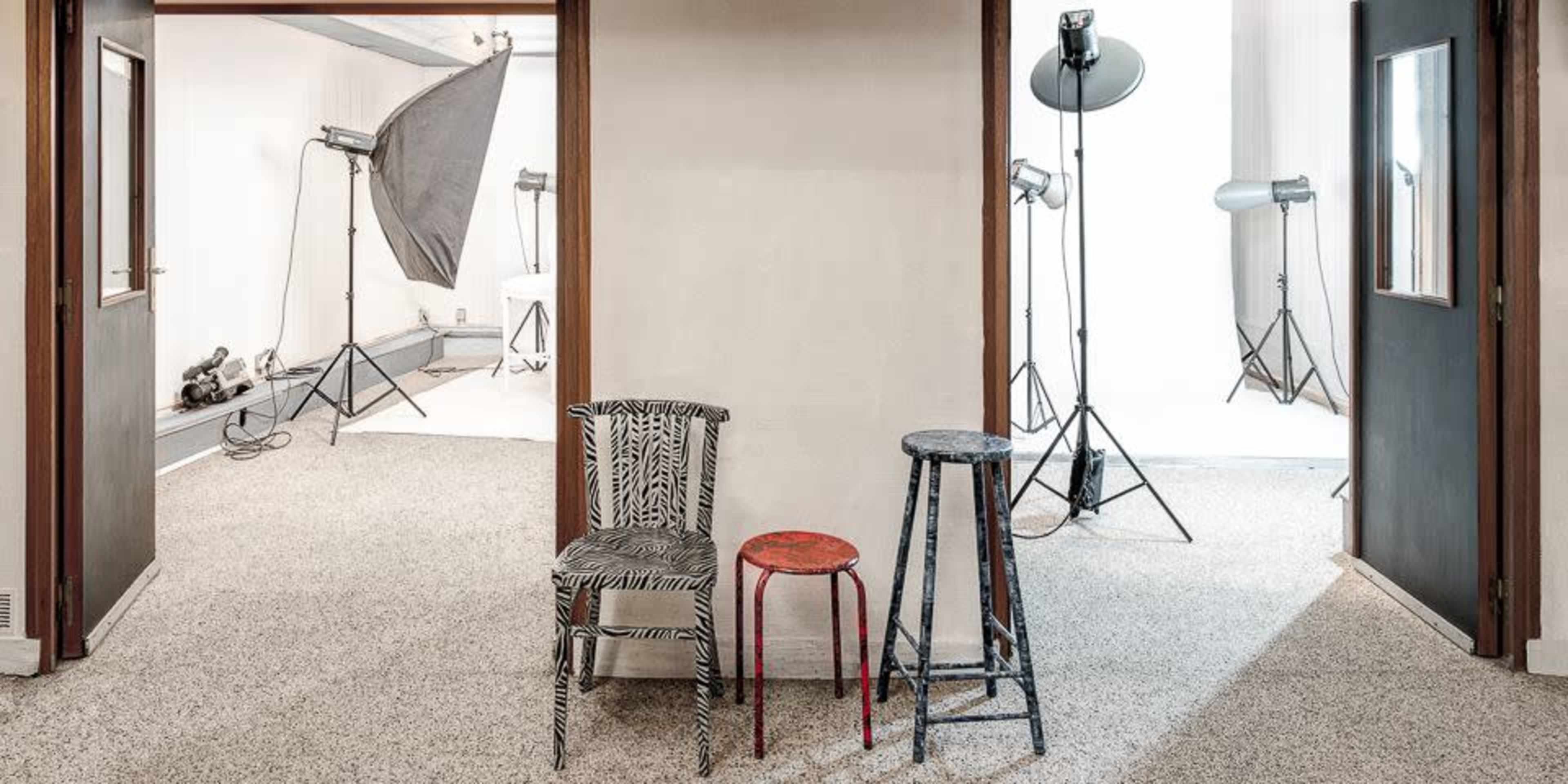 A hallway with two open doorways leading to photography studios, featuring various lighting equipment and three chairs with distinct designs placed in the center.