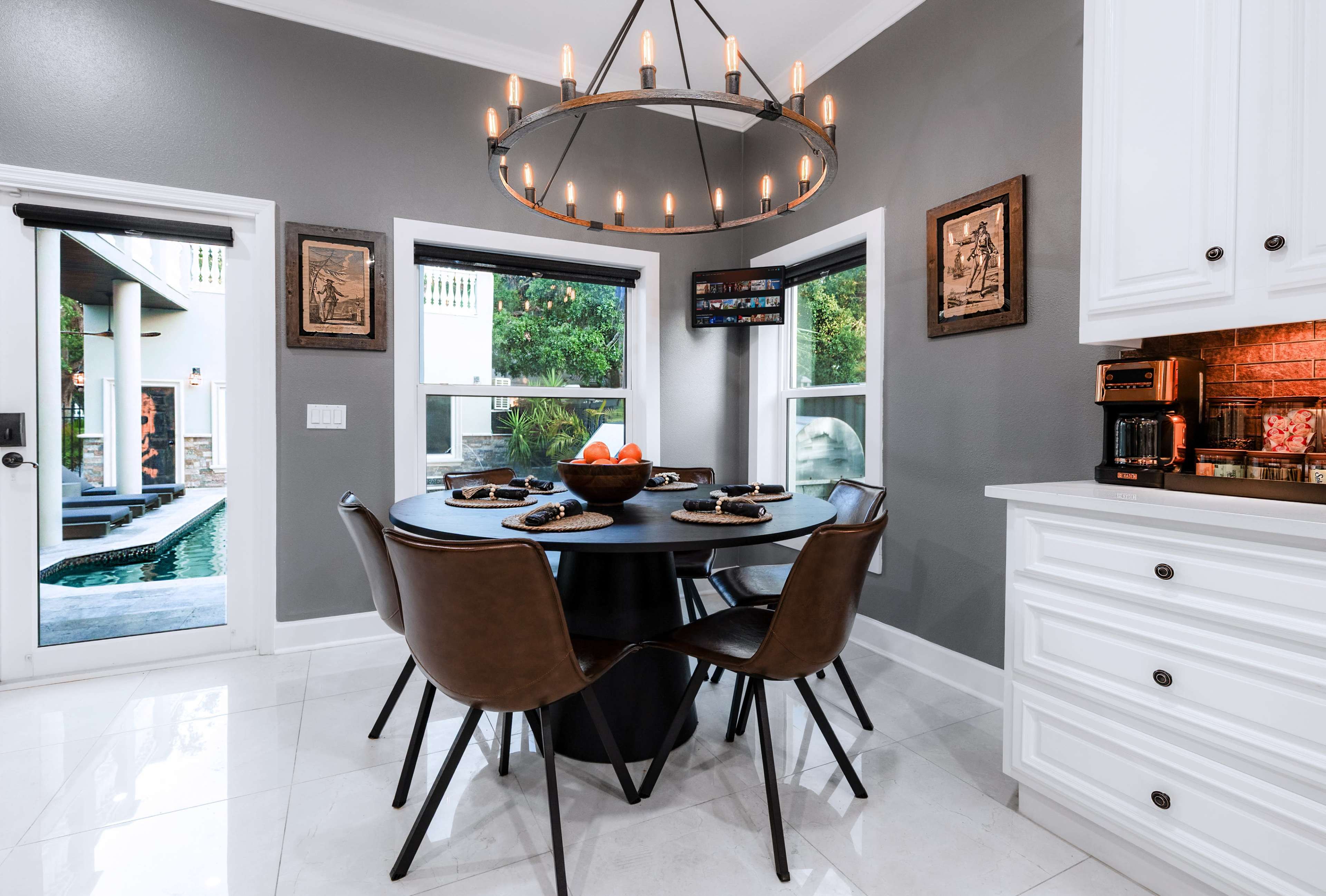 The image shows a modern kitchen with a round black dining table surrounded by brown chairs, set next to windows that overlook a pool area.