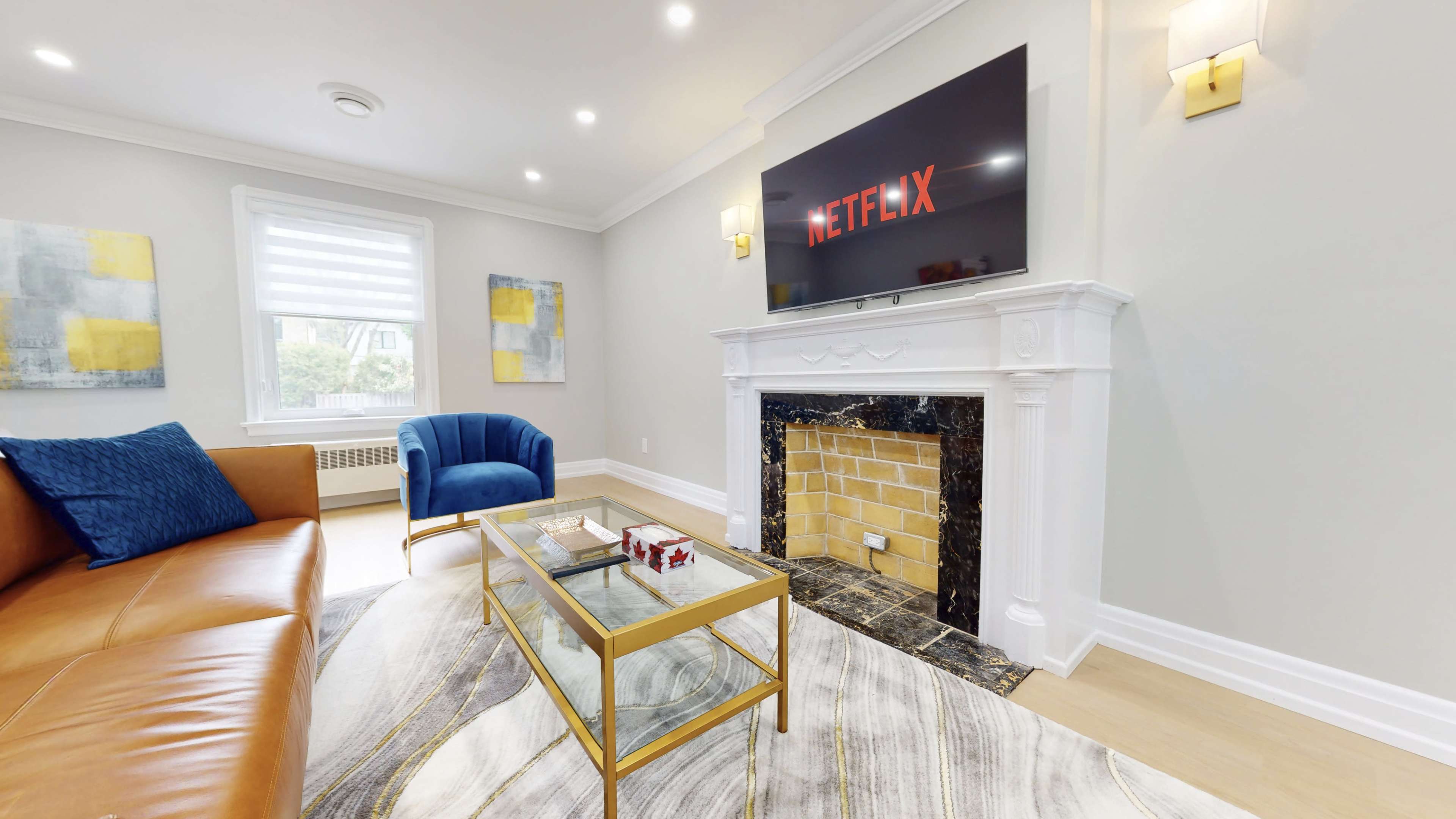 A modern living room with a blue armchair, a glass coffee table on a patterned carpet, and a television mounted above a white fireplace.