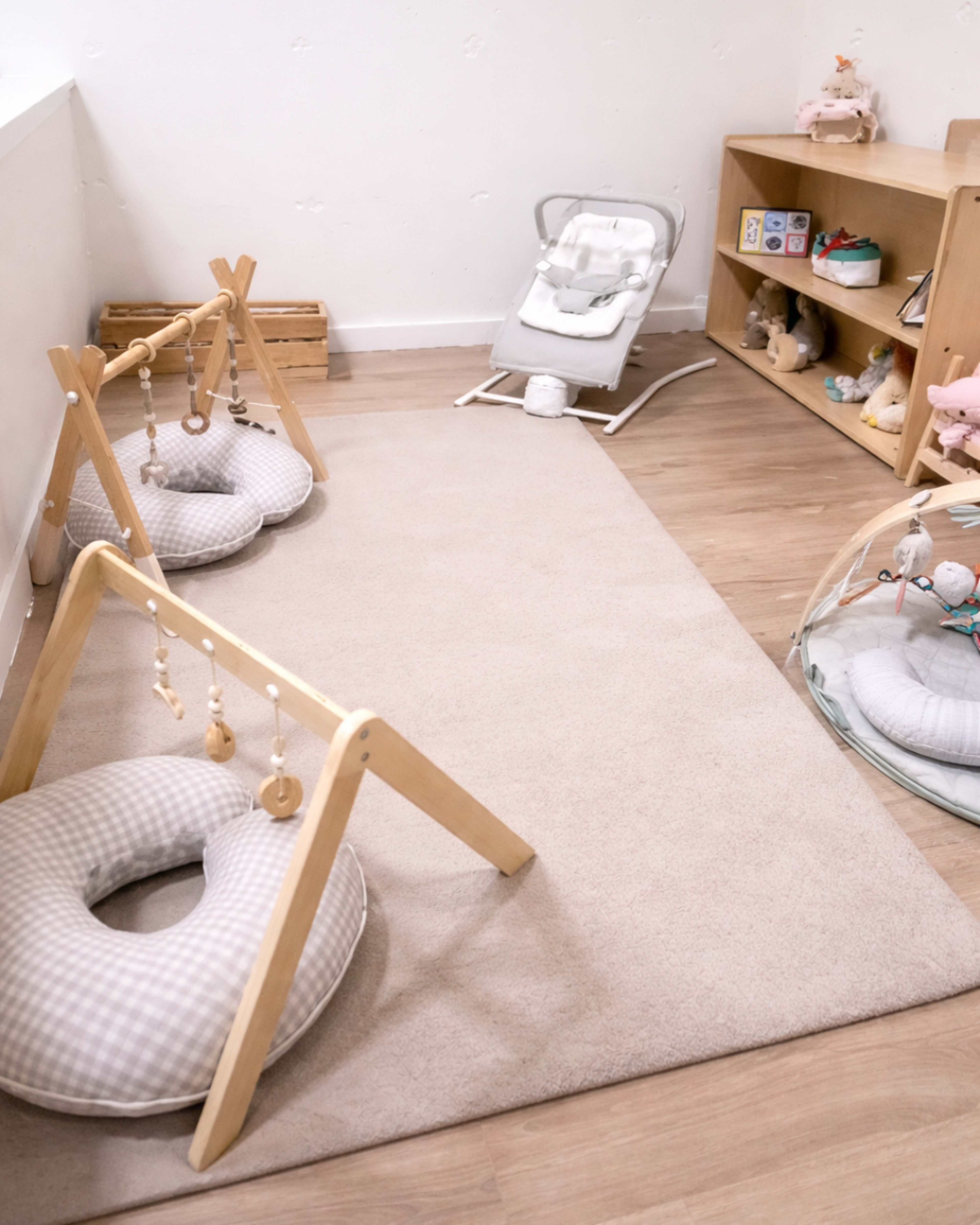 The image shows a softly lit nursery with a play mat, two wooden activity gyms, and a baby bouncer next to a shelf filled with toys.