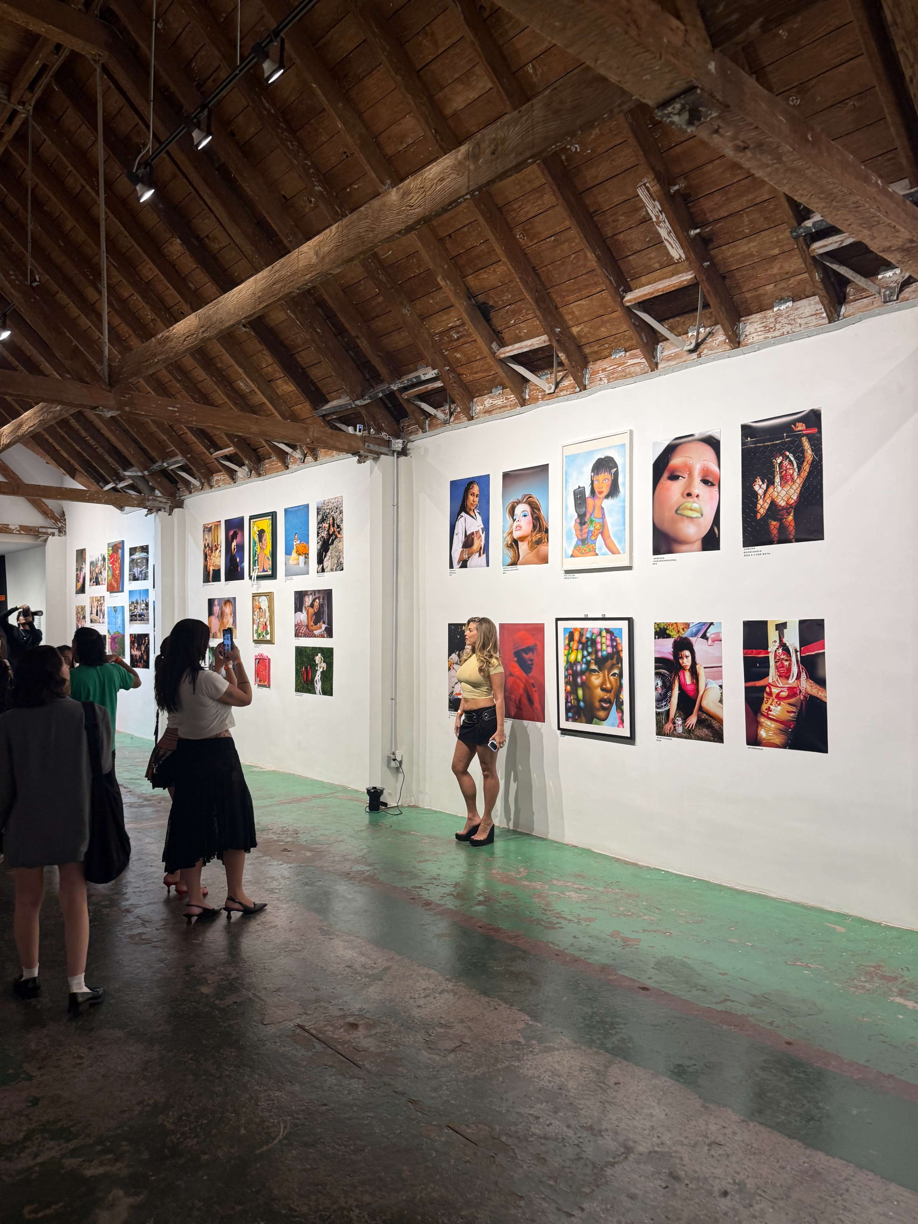 A group of people observes a gallery featuring a variety of colorful photographs displayed on white walls in a spacious, rustic room with wooden beams.