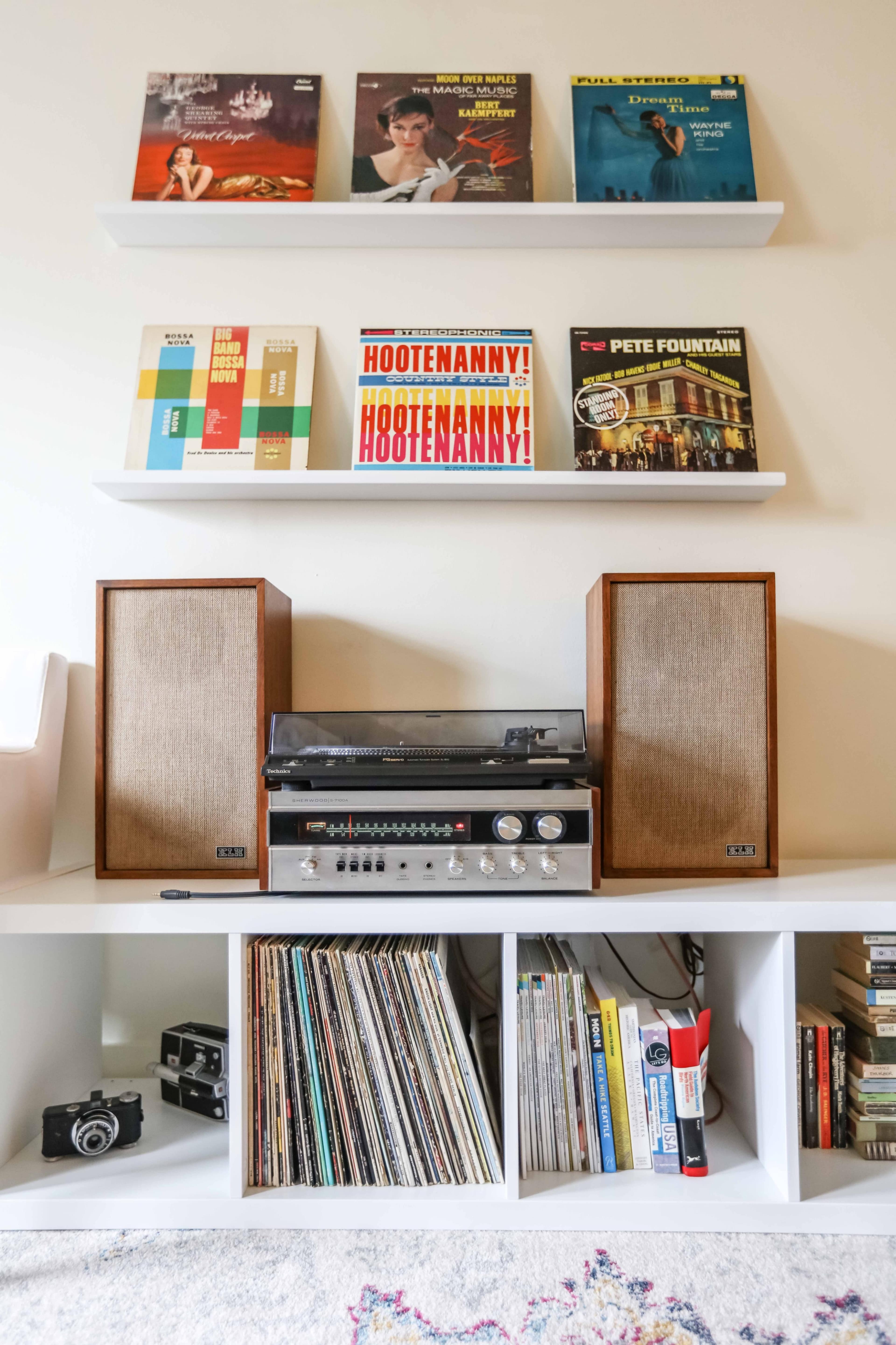 A vintage stereo system with wooden speakers is placed on a white shelf, surrounded by vinyl records and books.