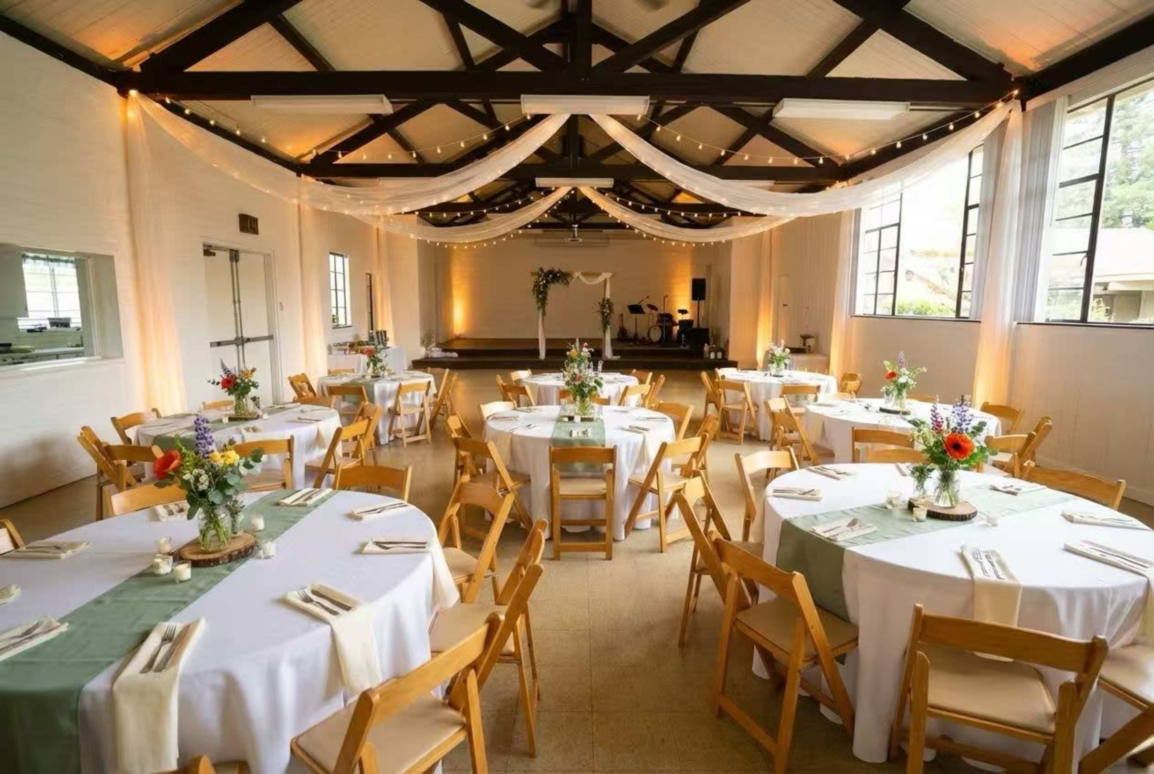The image shows a decorated indoor event space with round tables set for a celebration, featuring centerpieces of flowers and a stage at the front.