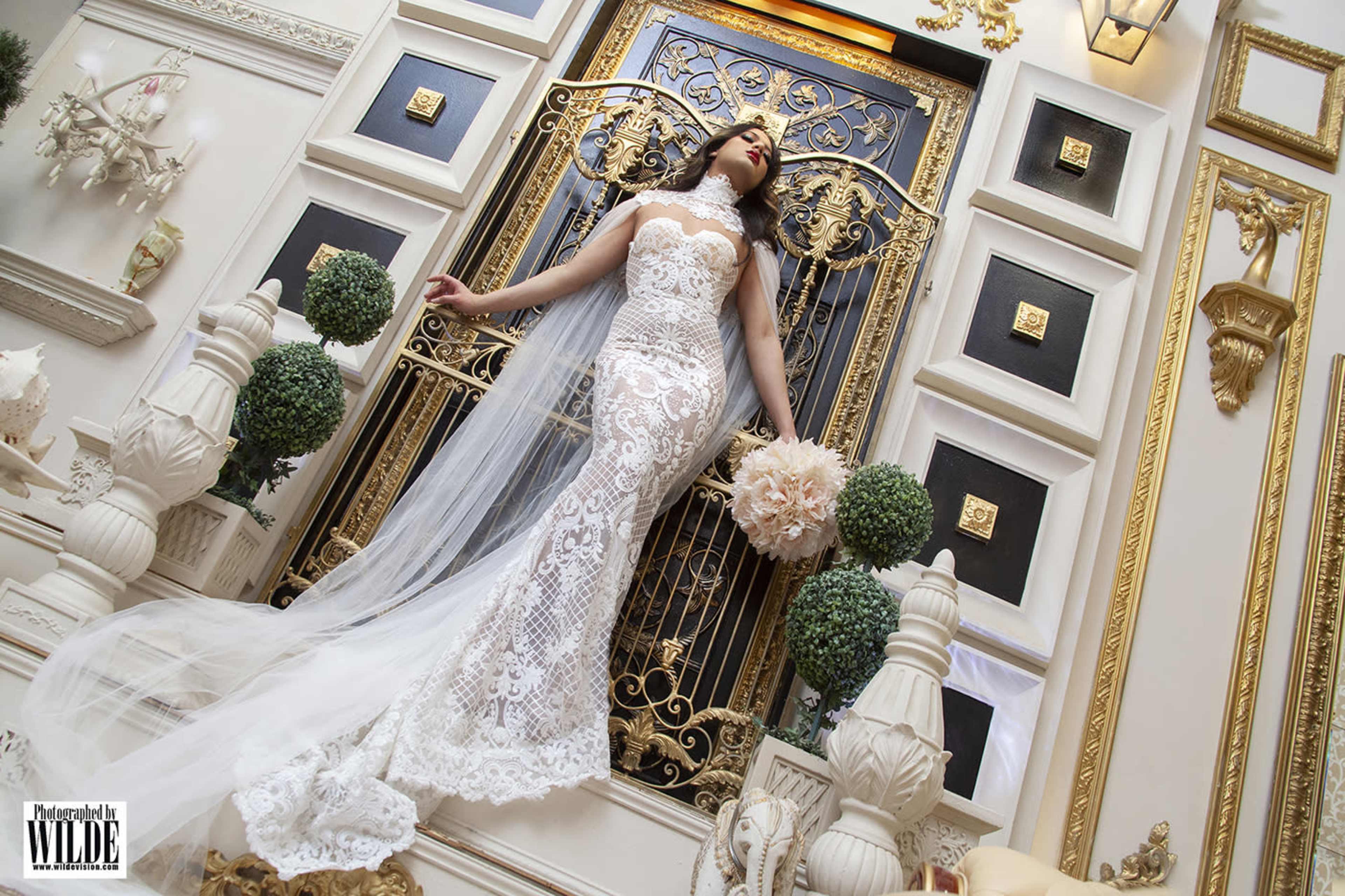 A woman in an intricate lace gown stands elegantly in front of a decorative golden door, holding a bouquet of flowers.