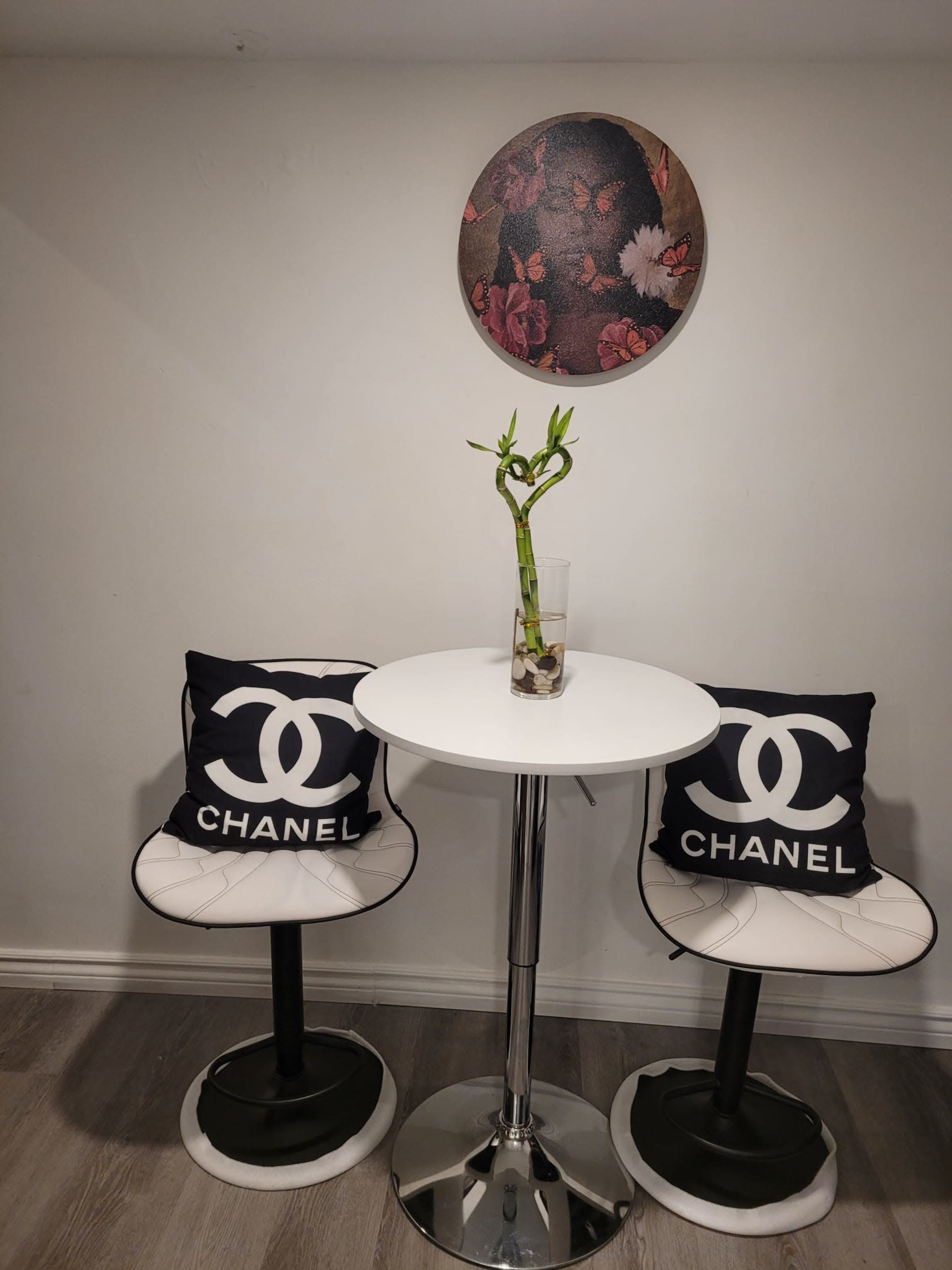 A small dining area features a round table with two Chanel-branded chairs, a vase with bamboo, and a floral wall decor above.