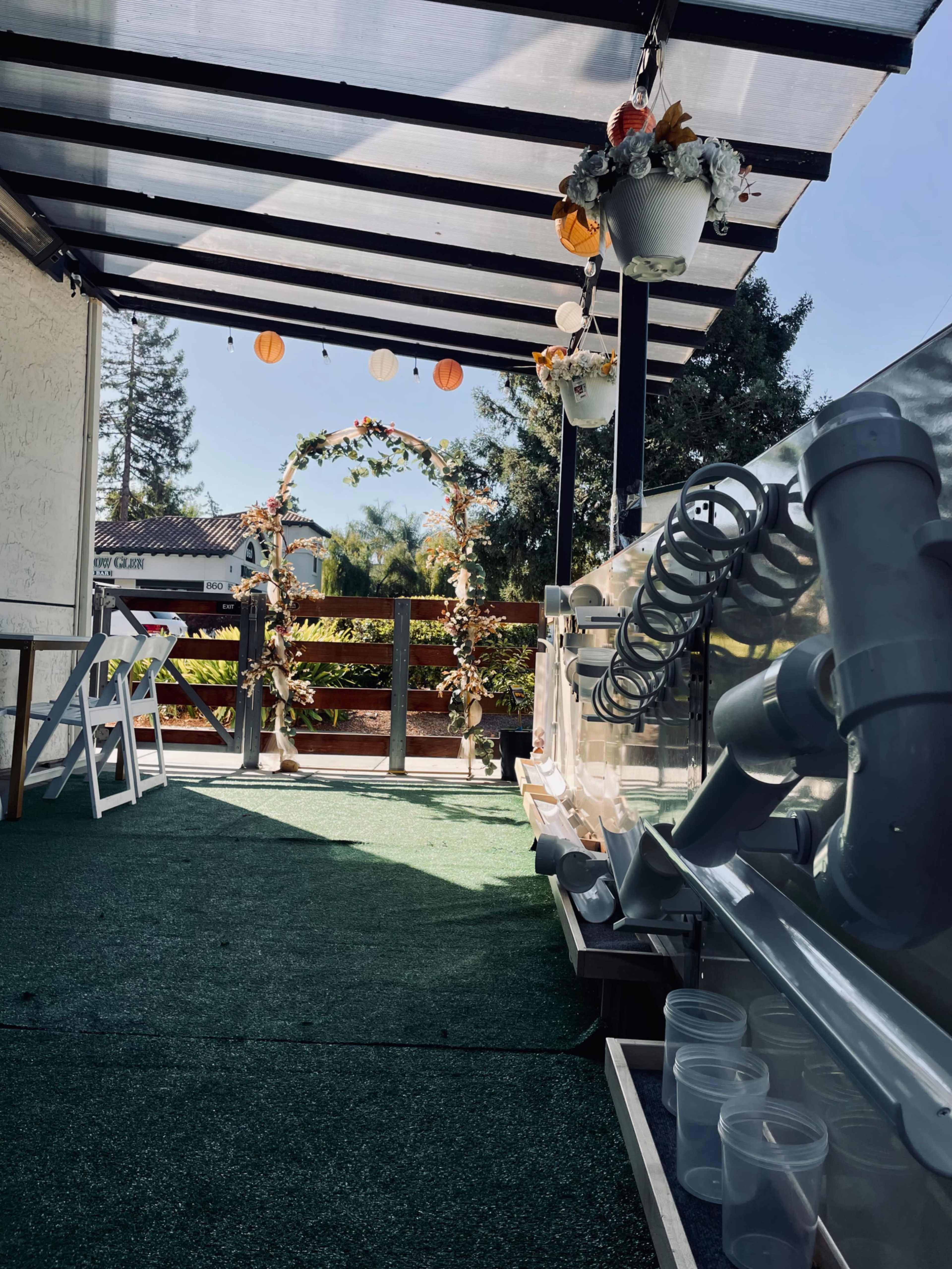 The image shows a decorated outdoor space with a green carpet, a floral arch, and a row of clear cups along a pipe structure.
