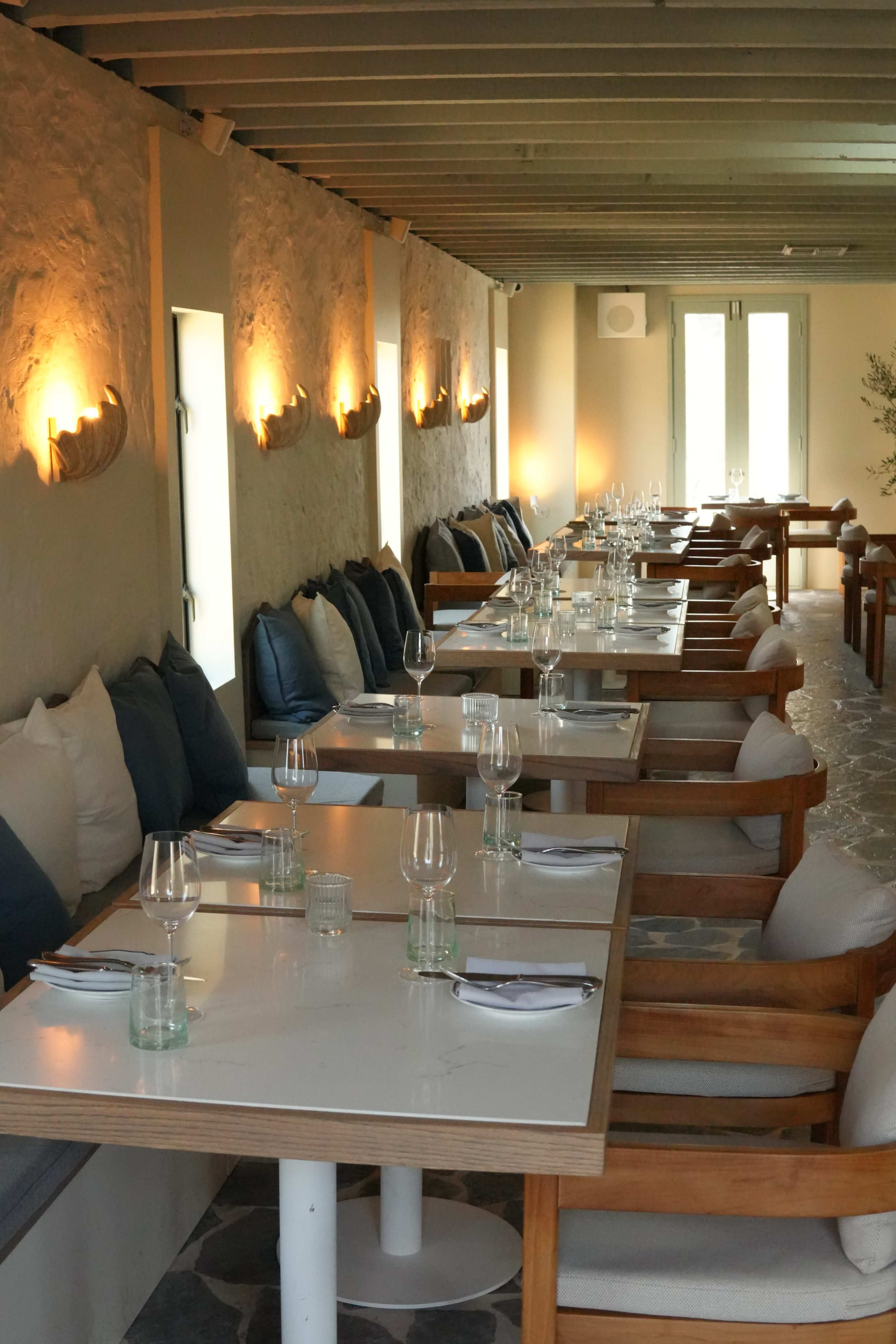 The image shows a modern restaurant interior with rows of tables set for dining, featuring wooden chairs and soft cushions against a textured wall.
