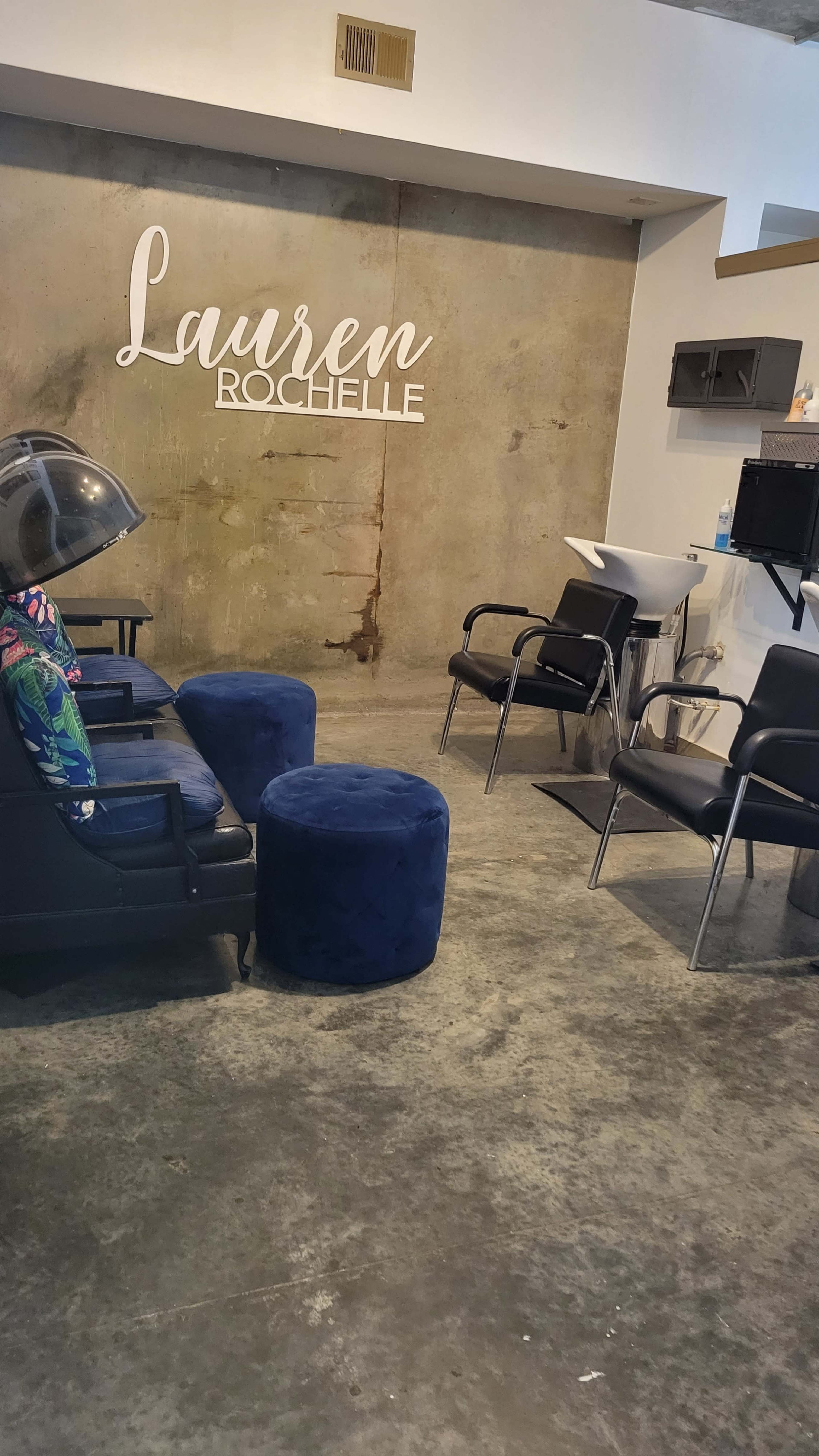 A hair salon interior featuring a dark chair with a hooded dryer, a round ottoman, and a reception area with a counter against a concrete wall that has the name "Lauren Rochelle" displayed.