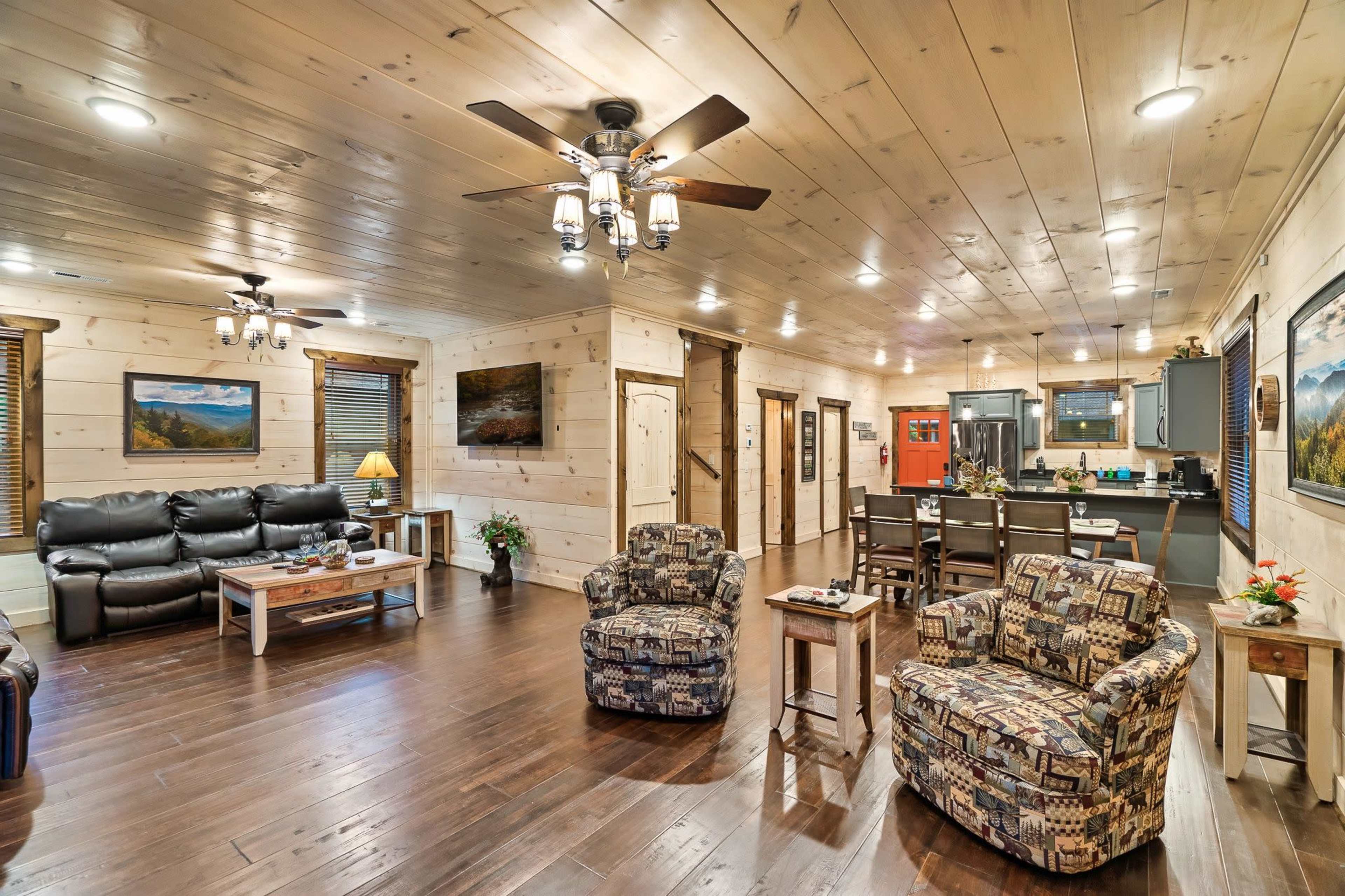 A spacious, modern living room with wooden walls and flooring, featuring a seating area with two patterned armchairs, a black leather sofa, and a dining area in the background.
