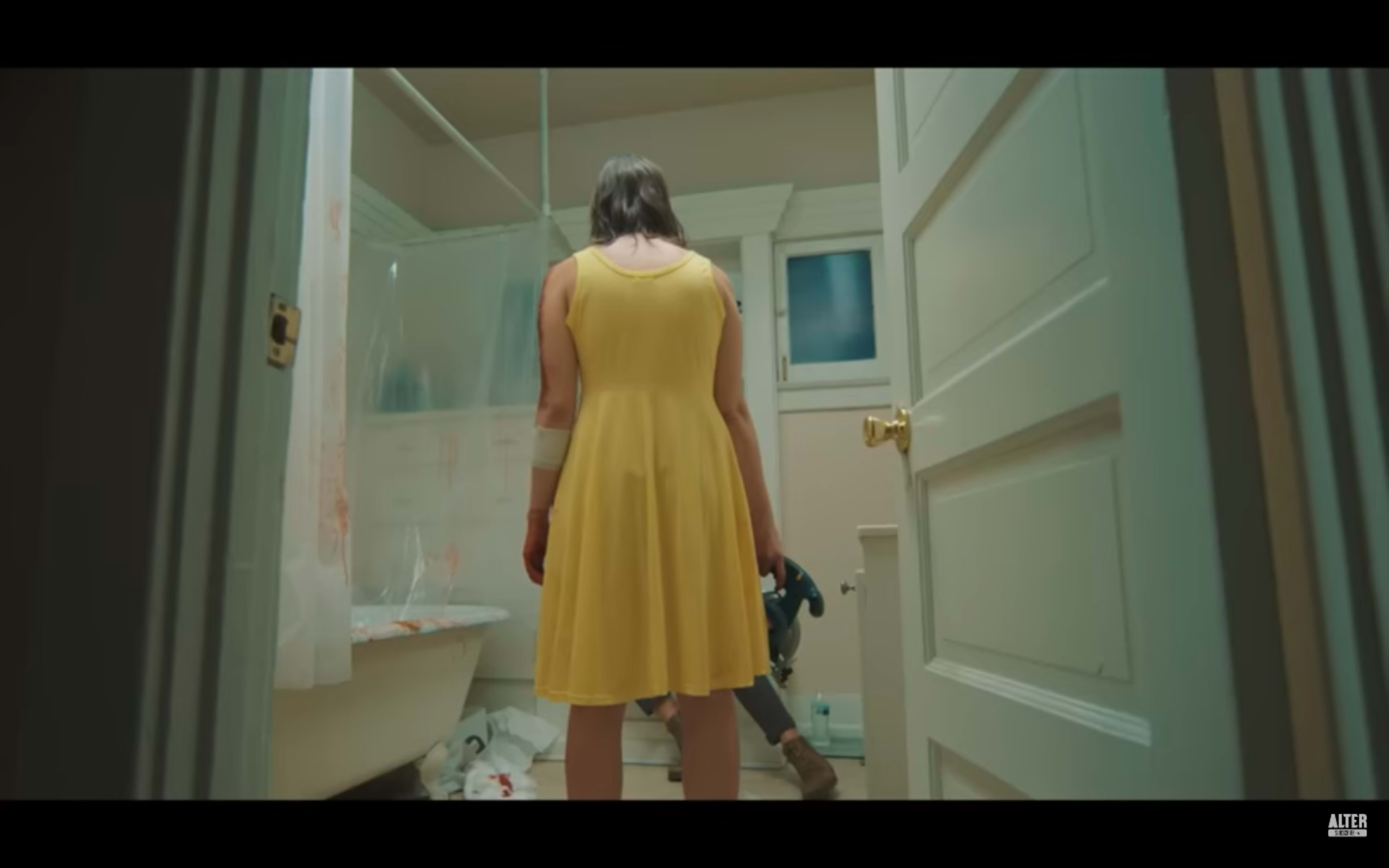 A person in a yellow dress stands in a bathroom, facing away and holding a power tool, with bloodstains visible on the walls and floor.