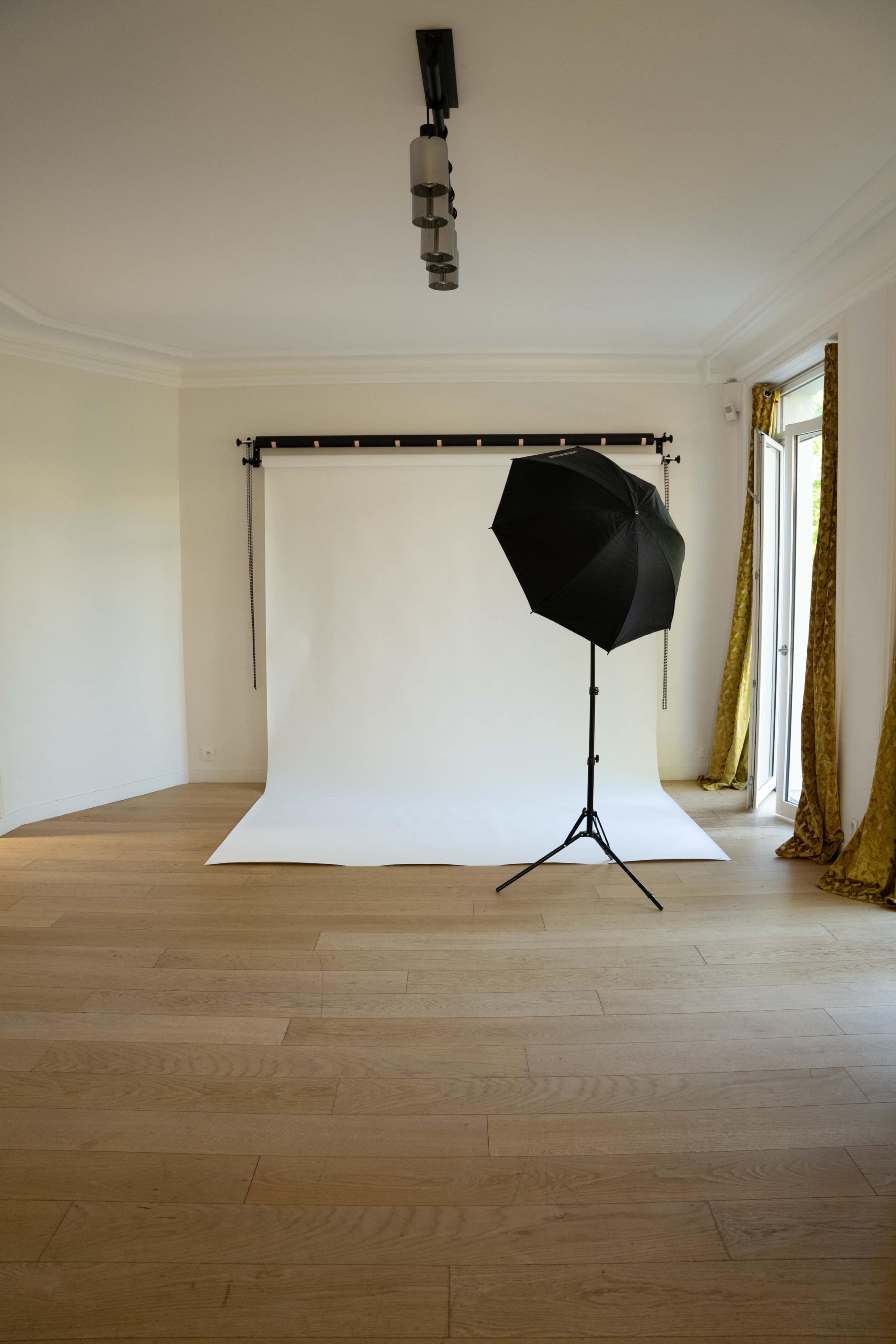 A photography studio features a white backdrop and an umbrella softbox on a stand, with wooden flooring and a window draped in yellow curtains.