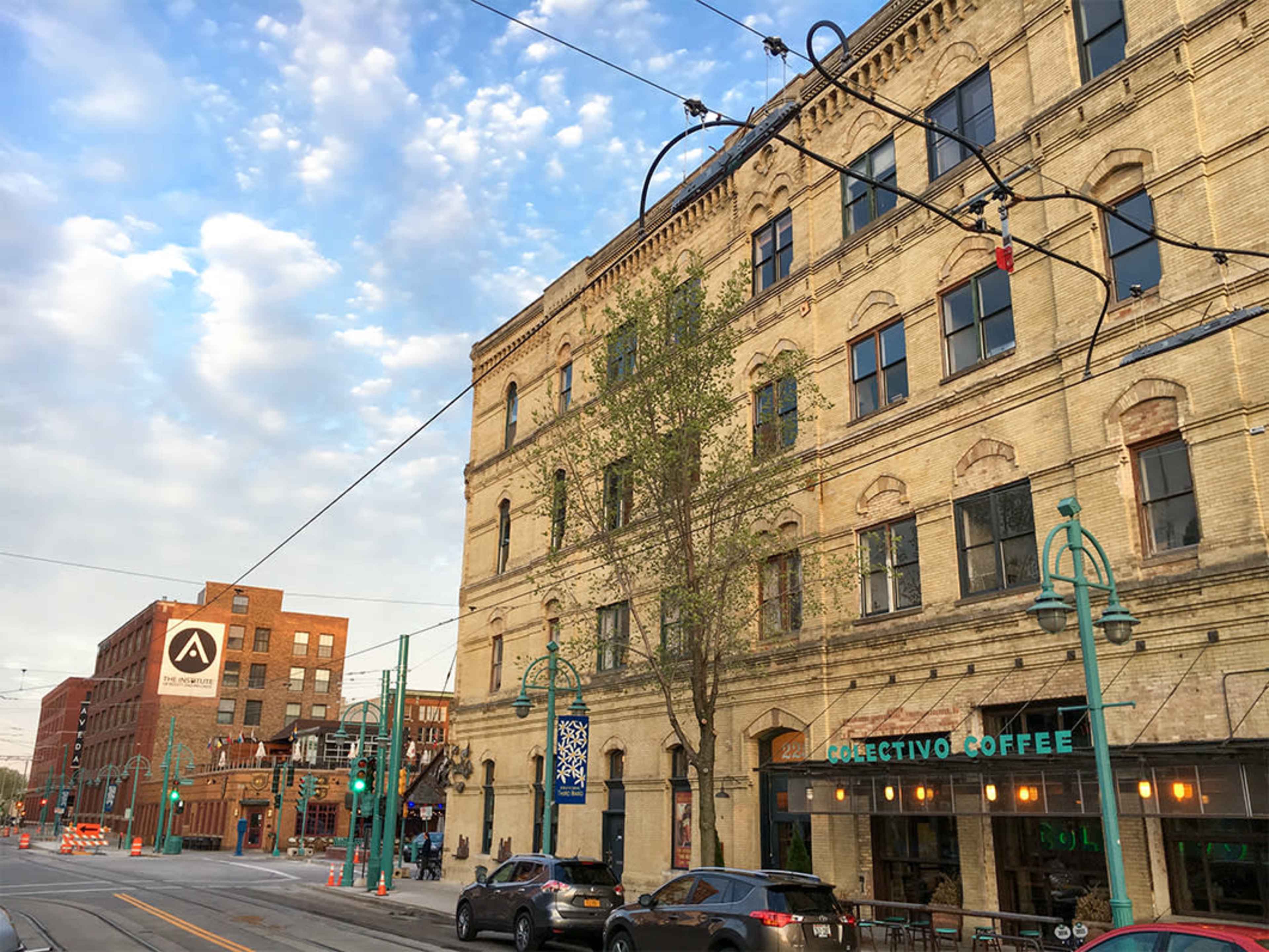 A street scene featuring a historic brick building with a sign for Colectivo Coffee, surrounded by power lines and trees under a partly cloudy sky.