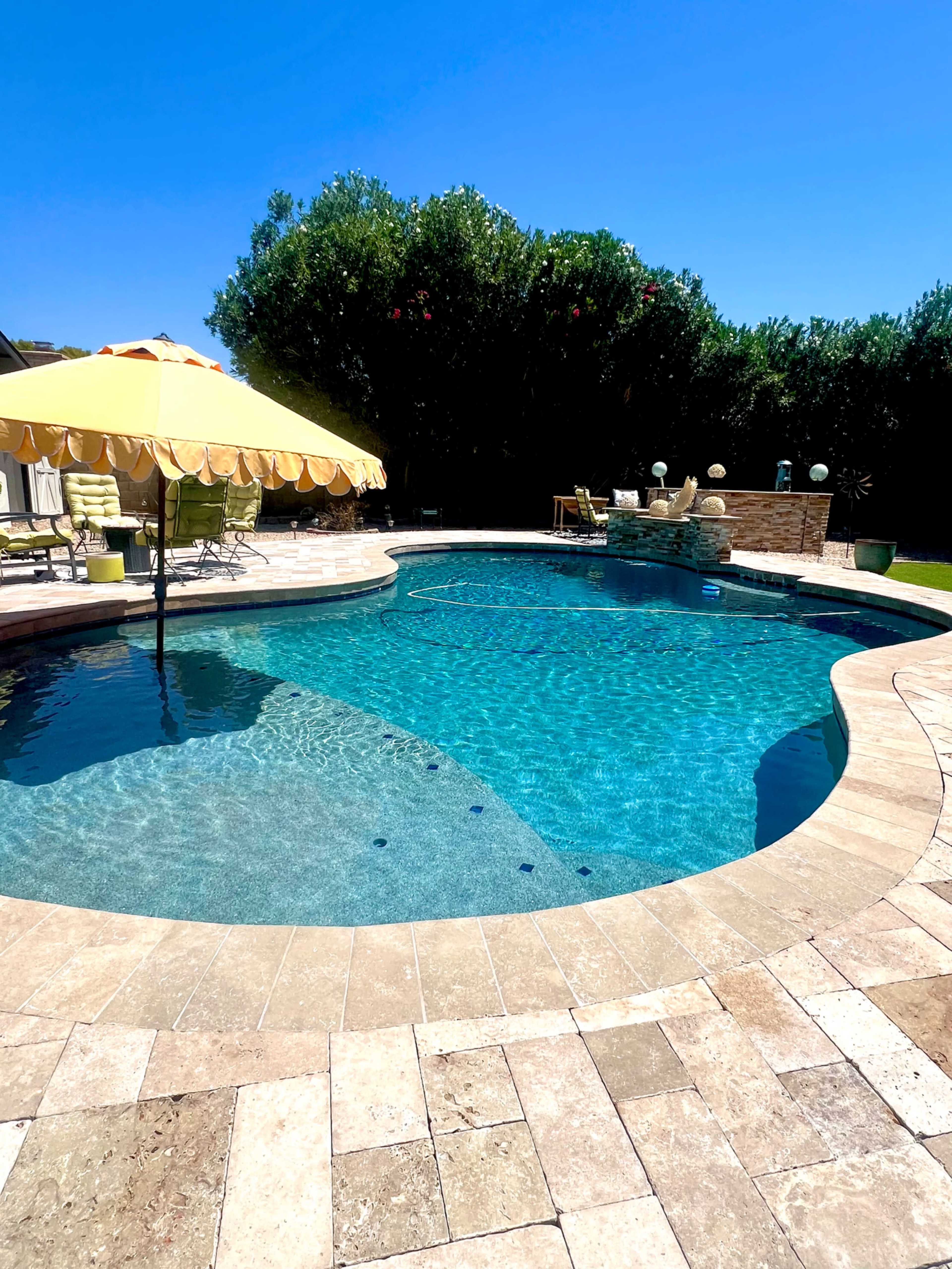 The image shows a clear swimming pool surrounded by a stone deck, with an umbrella and green lounge chairs in a well-maintained outdoor area.
