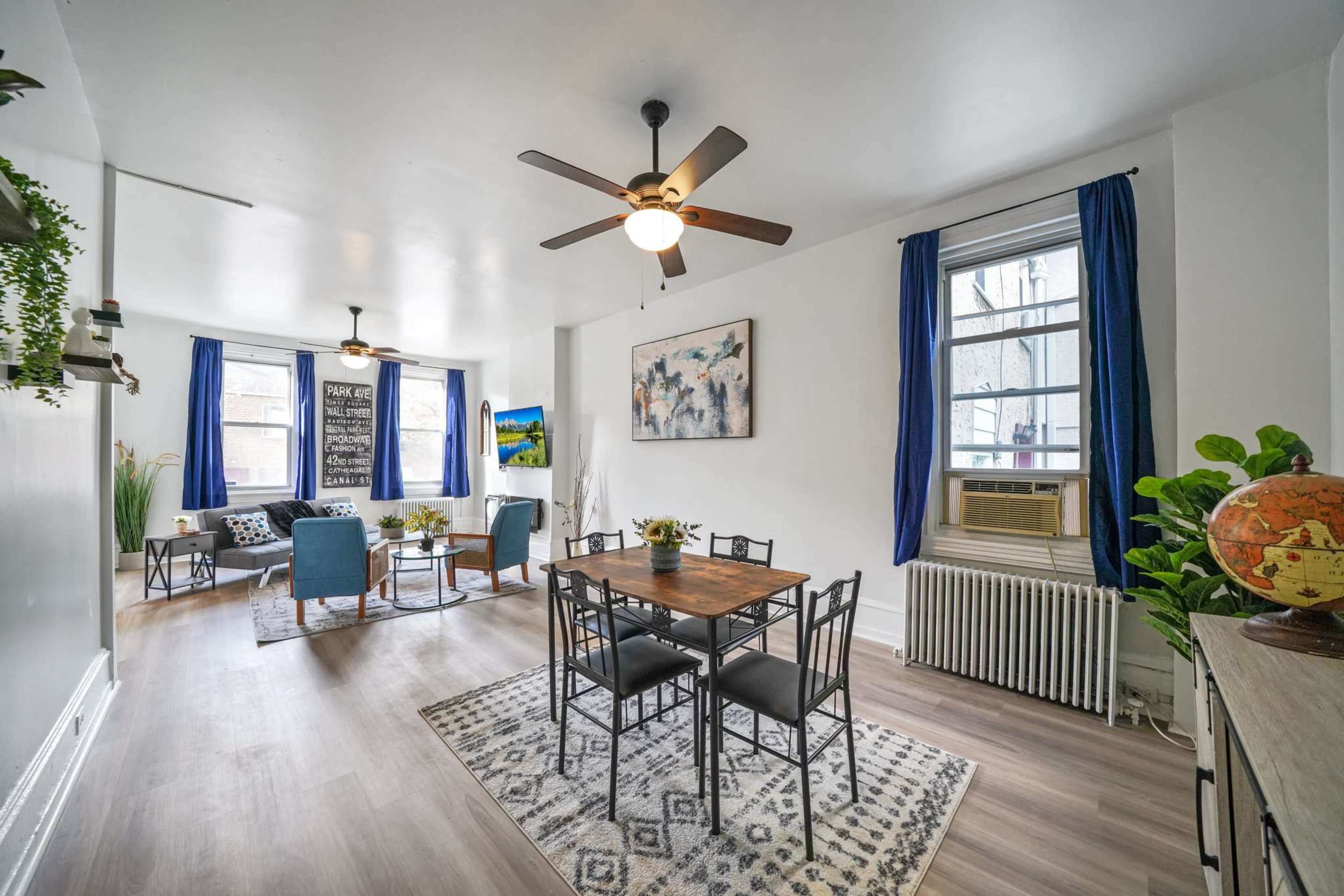 A bright, spacious living area featuring a dining table with six chairs, a seating arrangement with blue chairs, and large windows with blue curtains.
