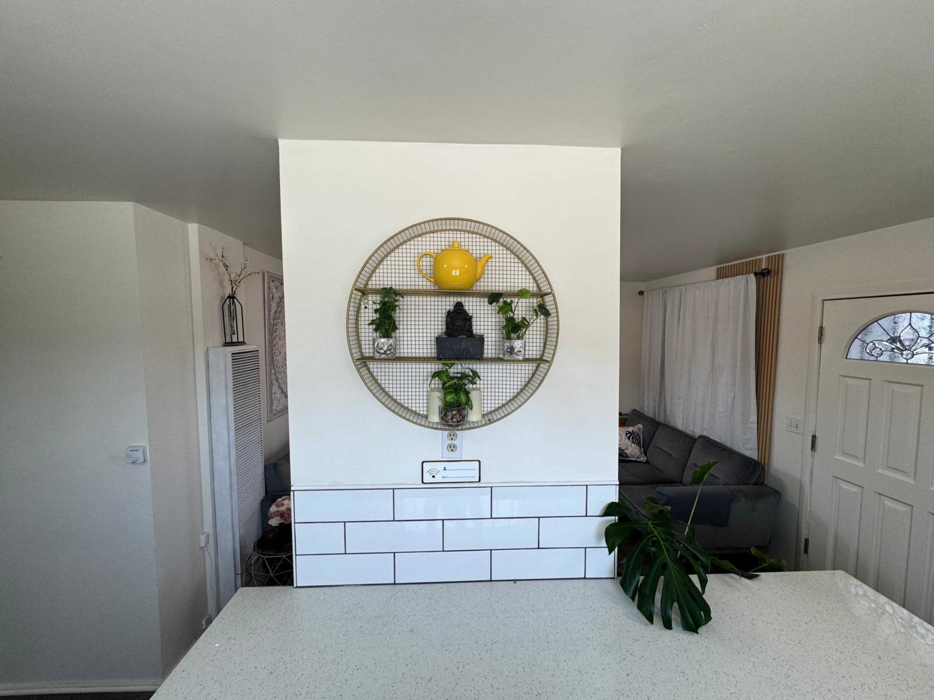 A round wall display with a yellow teapot and several plants is mounted on a white wall, separating the kitchen area from the living space in a bright room.