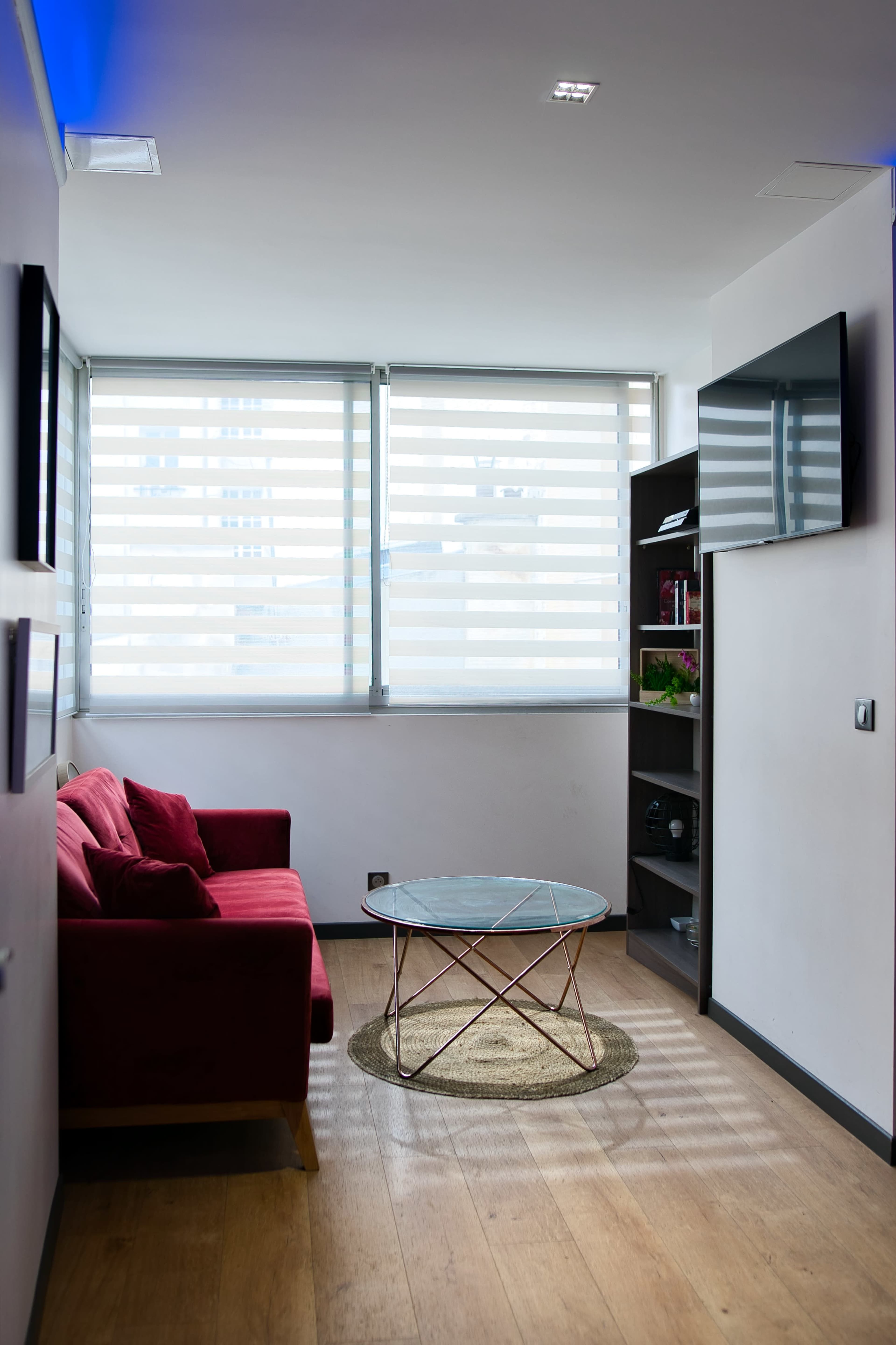 A modern living room features a red couch, a round coffee table, and large windows with horizontal blinds.