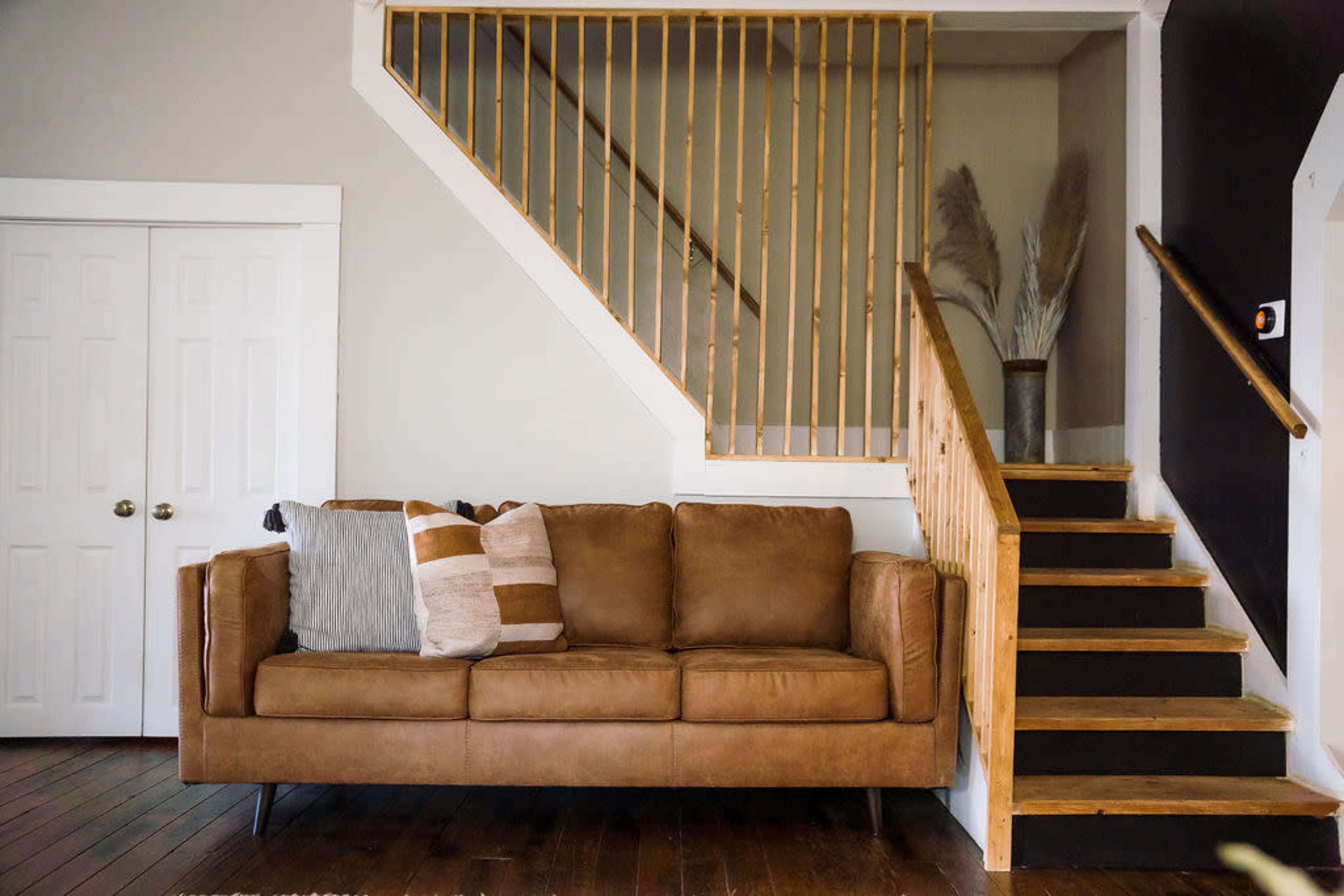 A brown upholstered sofa with decorative pillows is positioned next to a wooden staircase with a light-colored railing, set against a neutral wall in a well-lit room.
