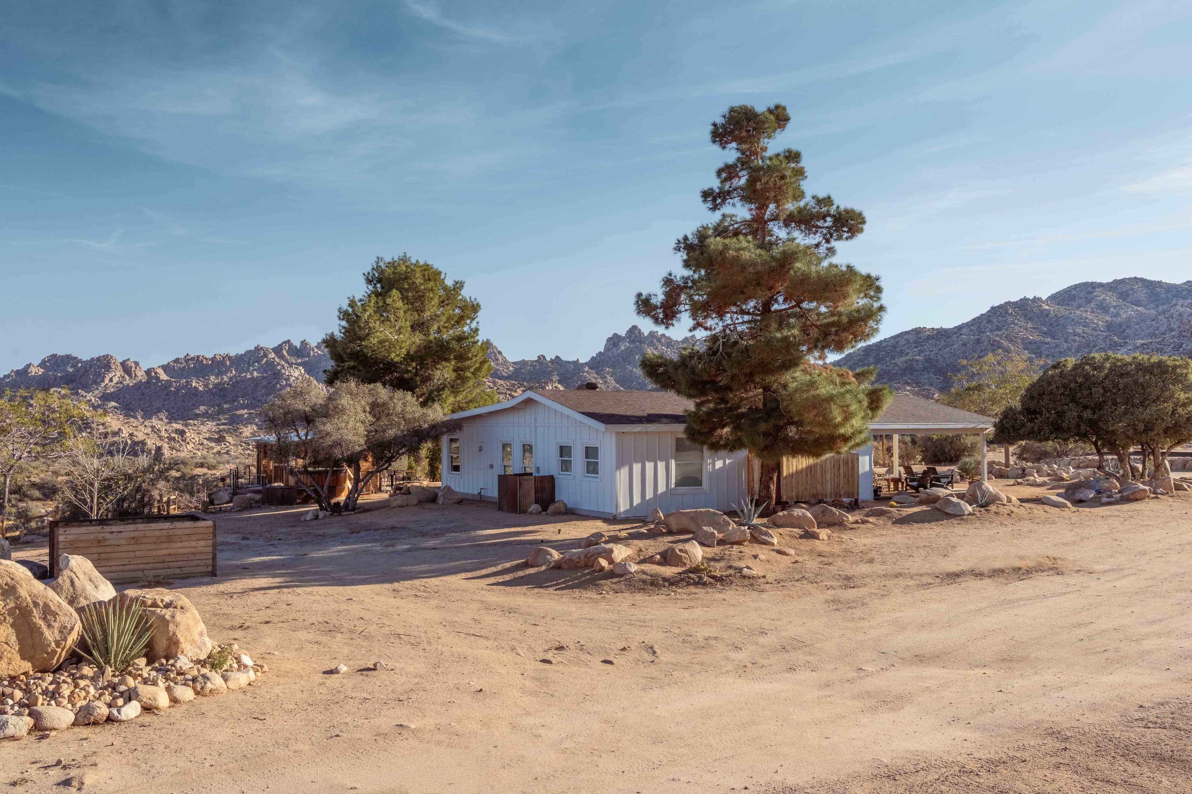A single-story white house is set against a backdrop of rocky mountains and desert vegetation in a remote area.