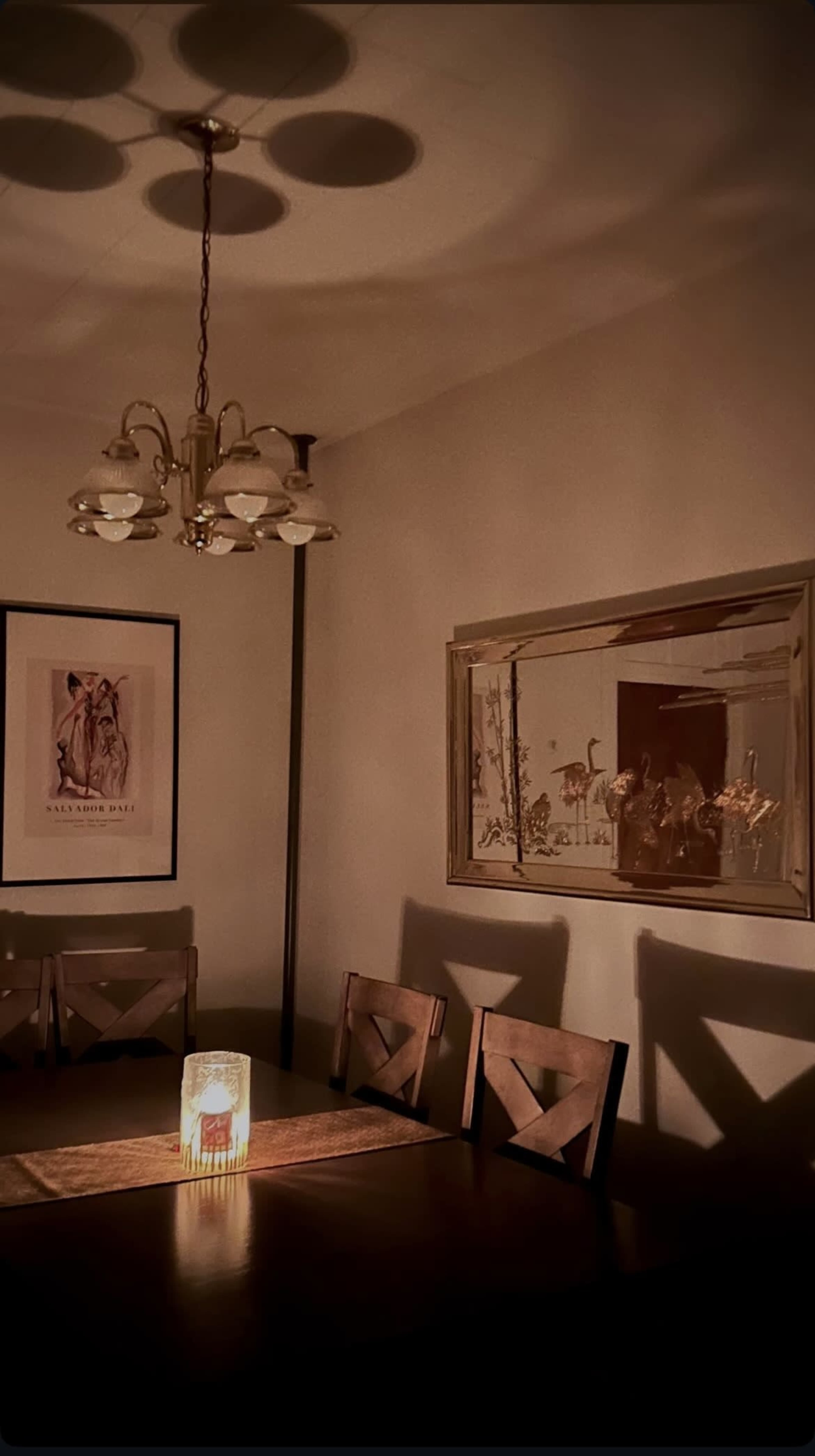 A dimly lit dining room features a table with a candle at its center, surrounded by wooden chairs and framed artwork on the walls.