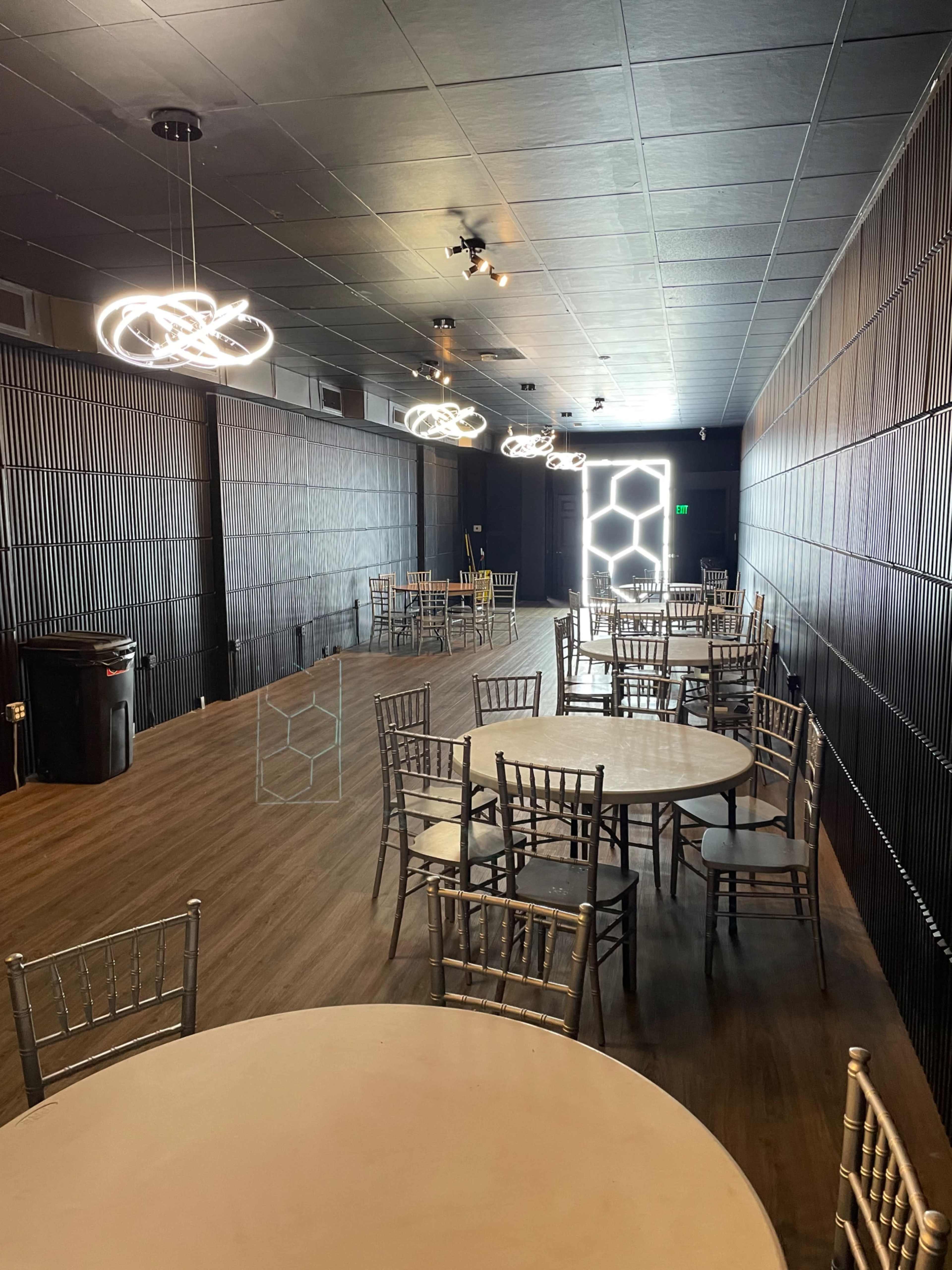 A spacious indoor venue with several round tables and metallic chairs, illuminated by modern light fixtures, leading to a doorway that features a geometric design.