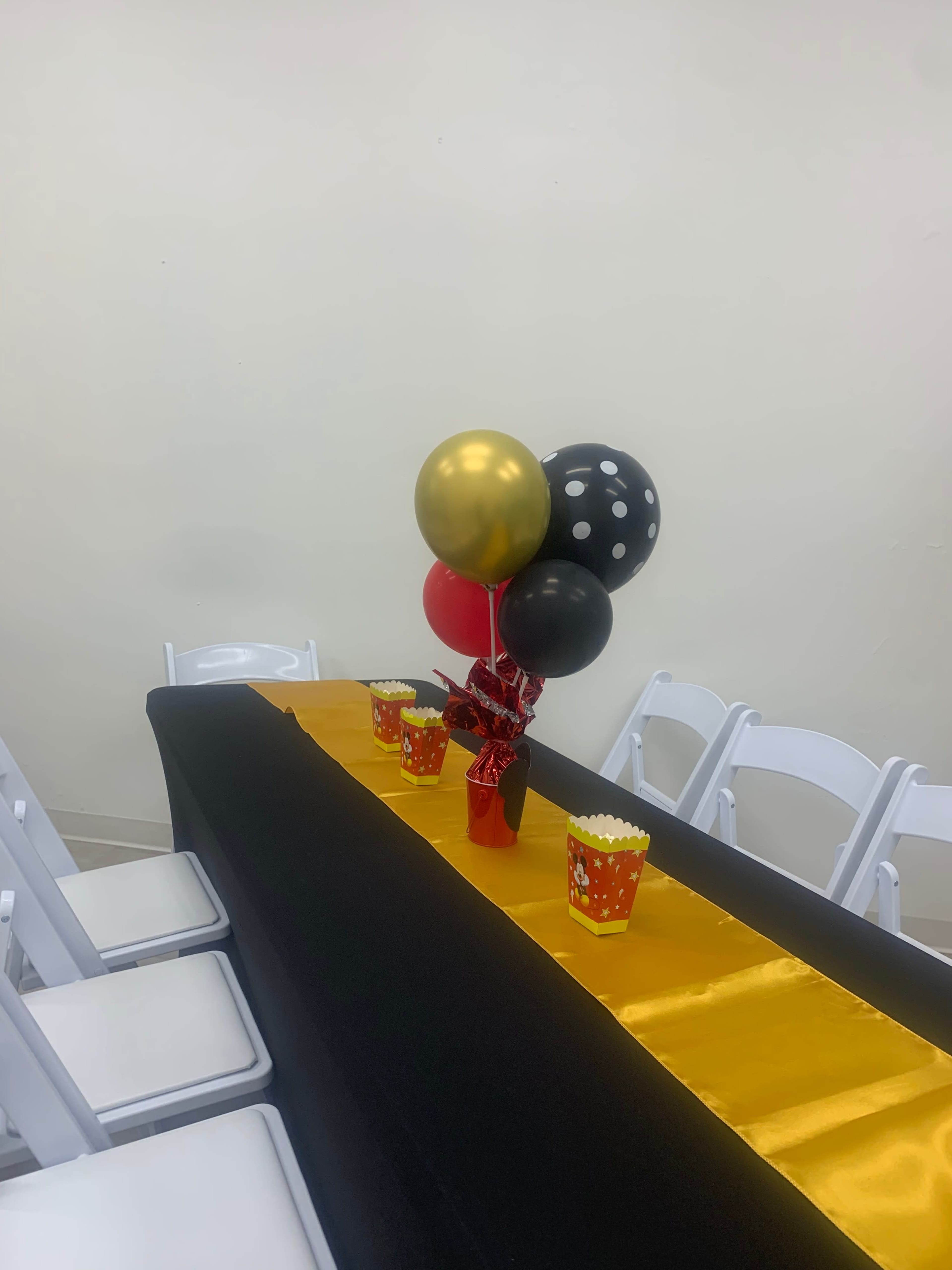 A table is set with a black tablecloth, a gold runner, popcorn containers, and balloons in black, gold, and red.