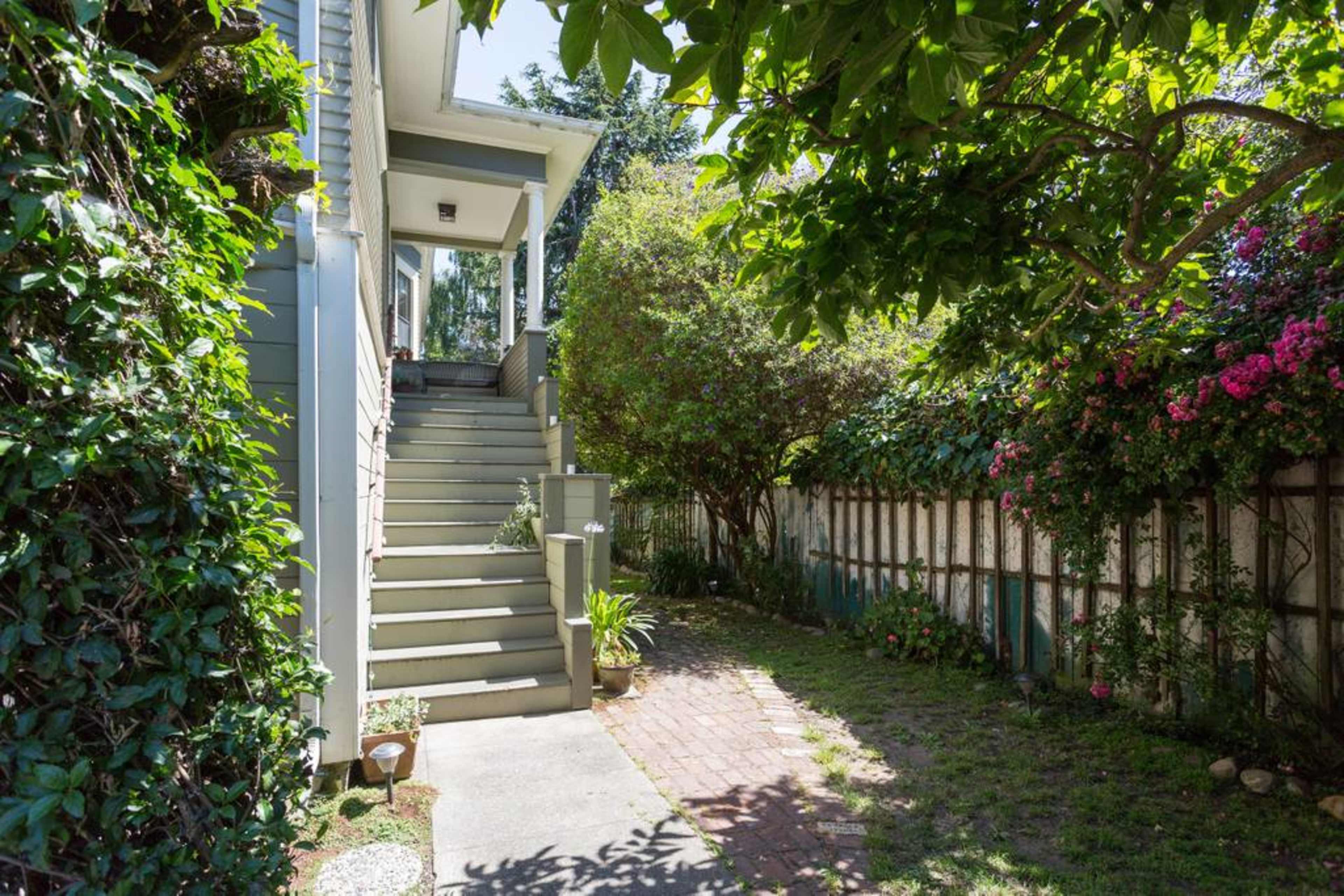 A pathway leads to a set of stairs by a house, flanked by greenery and flowering plants on both sides.