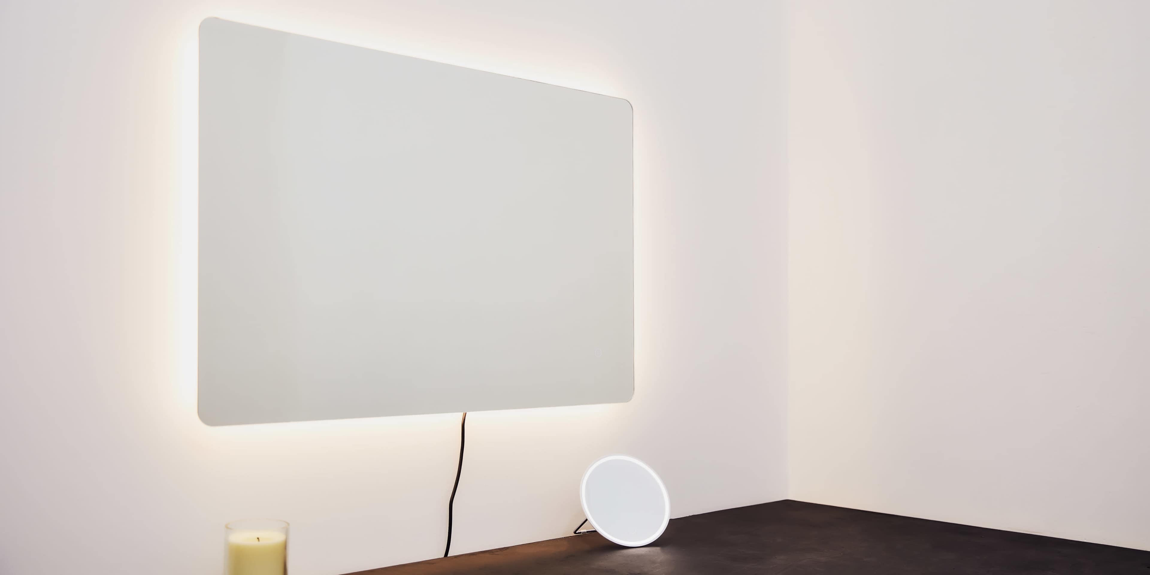 A wall-mounted mirror with backlighting is positioned above a small round mirror on a black surface in a softly lit room.