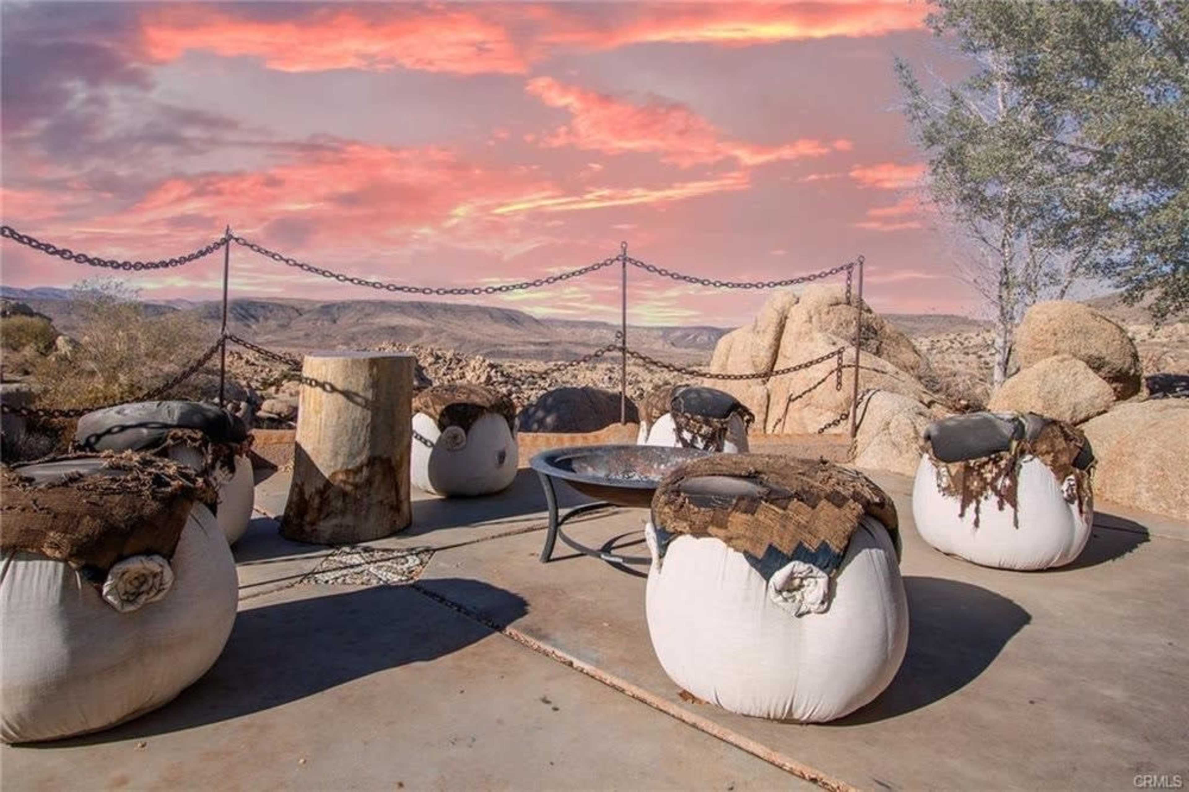 The image shows a cozy seating area with round chairs and a fire pit, set against a rugged desert landscape under a colorful sunset.
