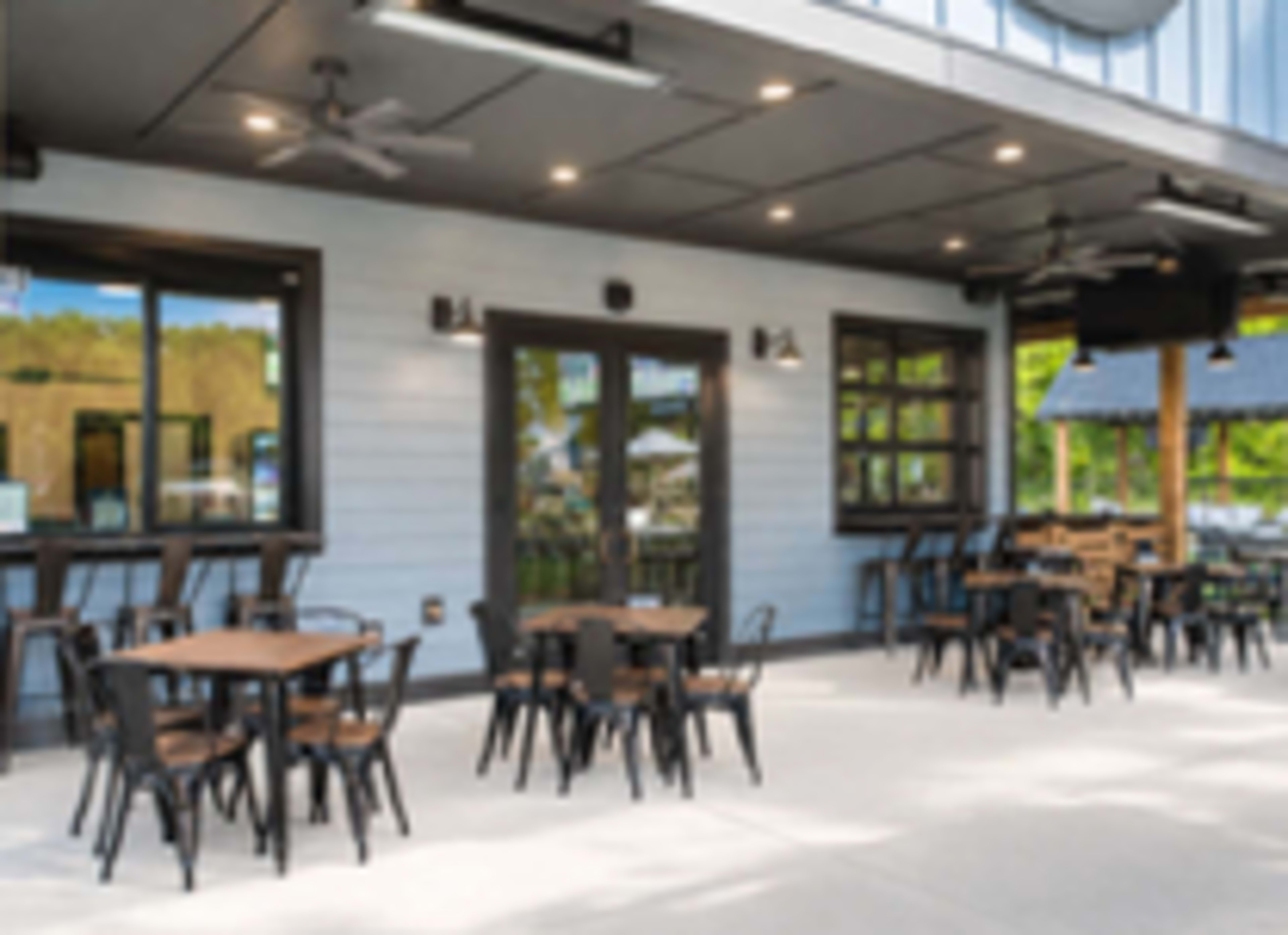 The image shows an outdoor dining area featuring several wooden tables and black metal chairs, with a blue wooden building in the background.