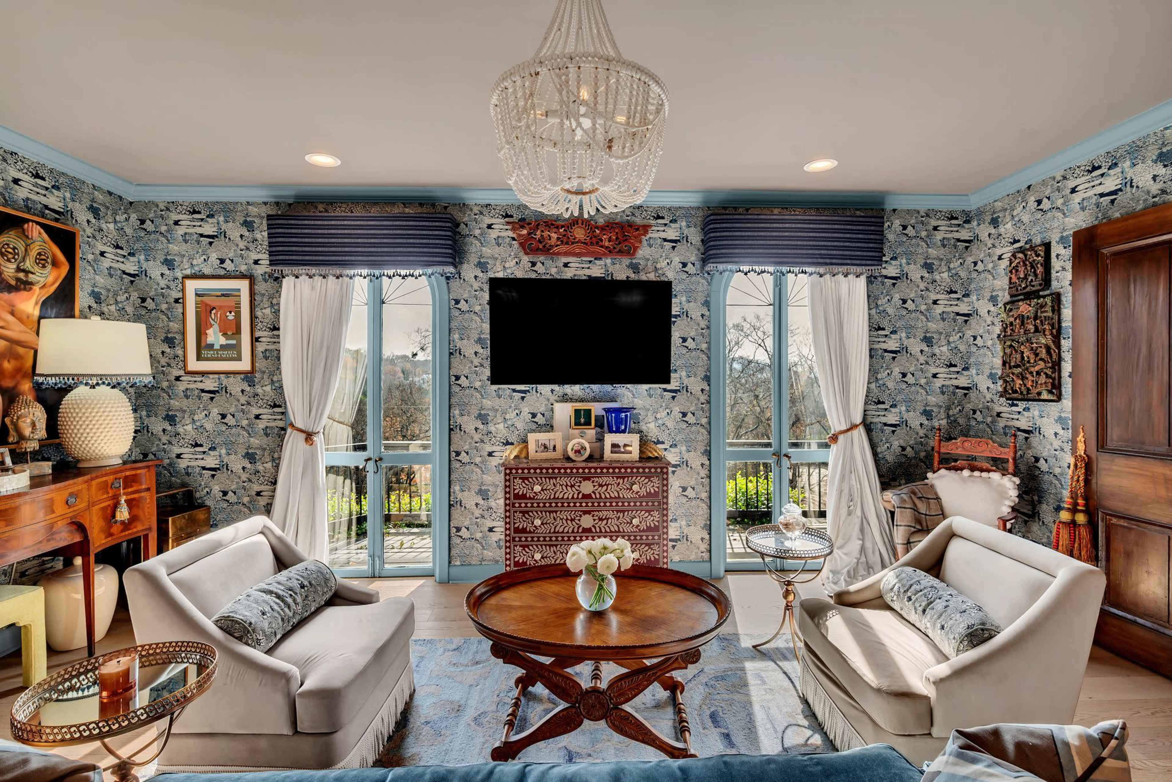 The image shows a living room with two white armchairs, a round wooden table, a patterned blue wallpaper, and a large wall-mounted television above a decorative cabinet.