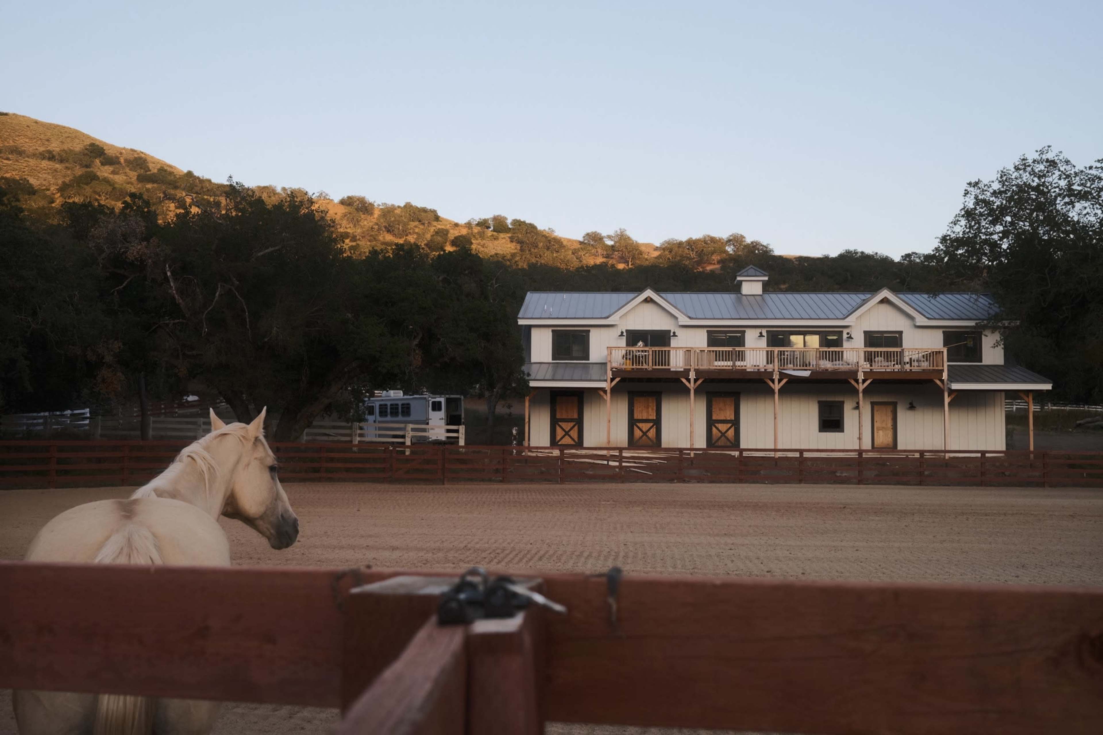 A horse stands near a wooden fence while a two-story stable is visible in the background against a hillside.