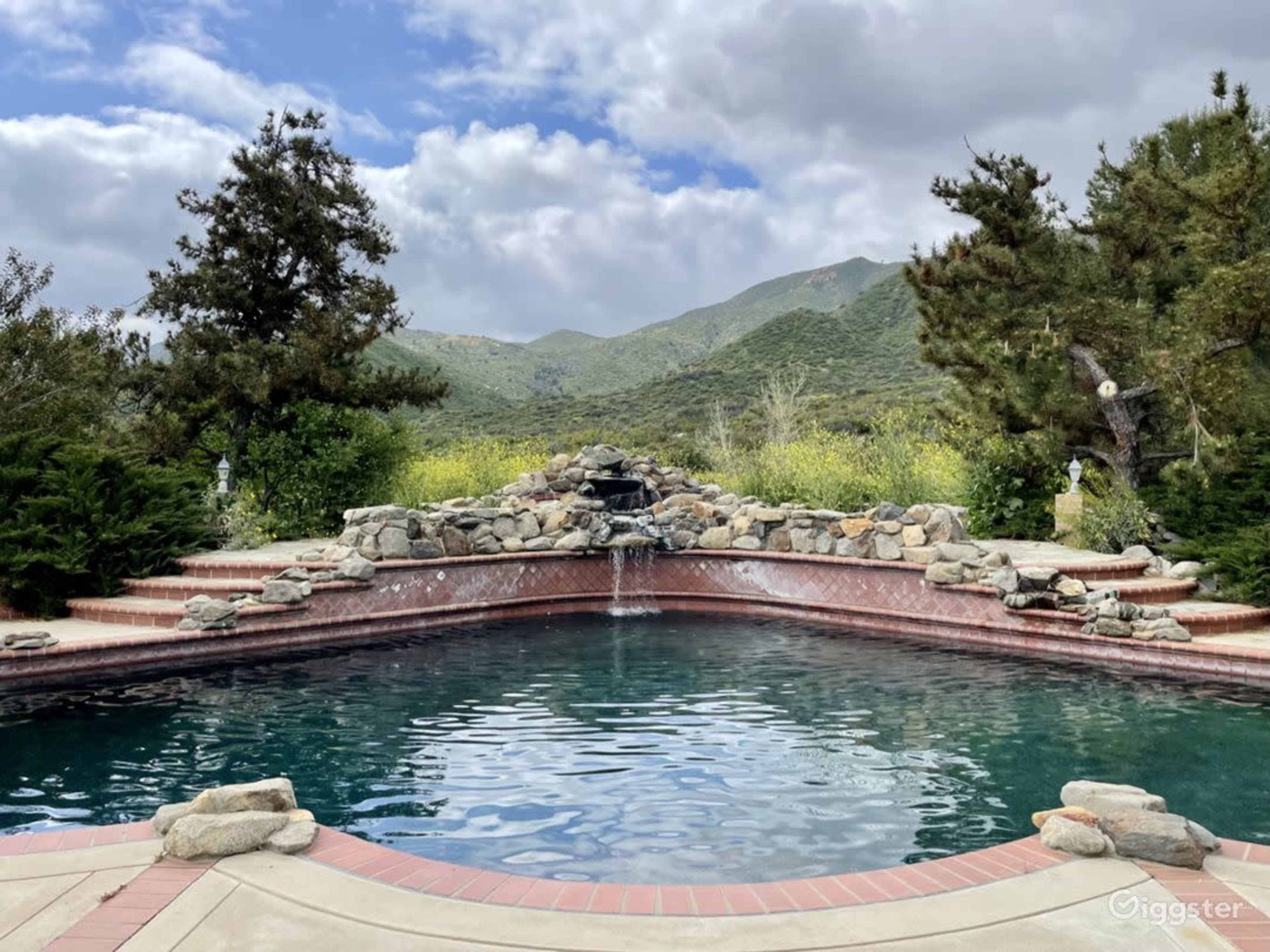 The image depicts a stone-lined swimming pool with a waterfall and mountain scenery in the background under a partly cloudy sky.