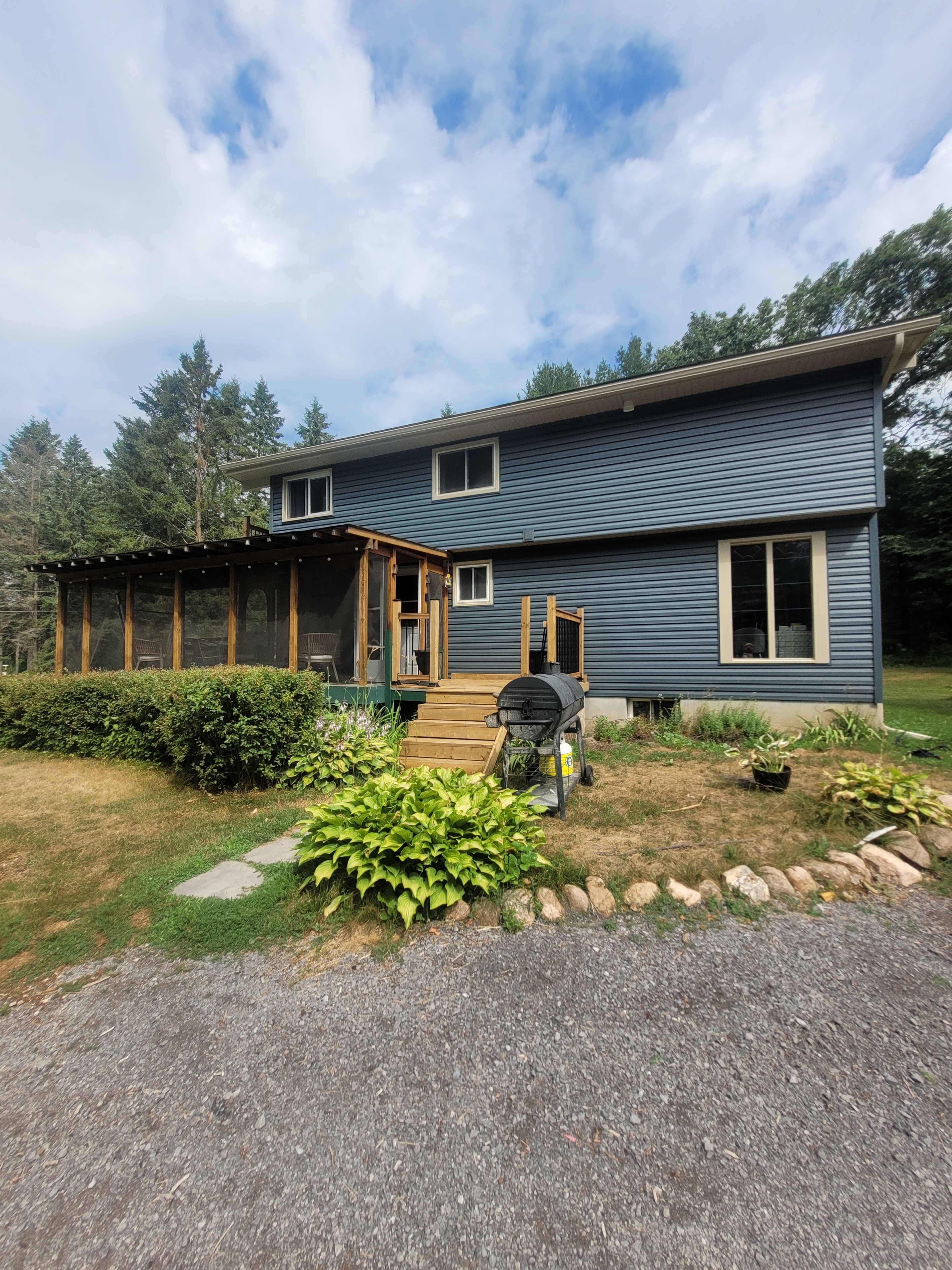 The image shows a two-story blue house with a wooden deck, surrounded by a landscaped yard and gravel driveway.