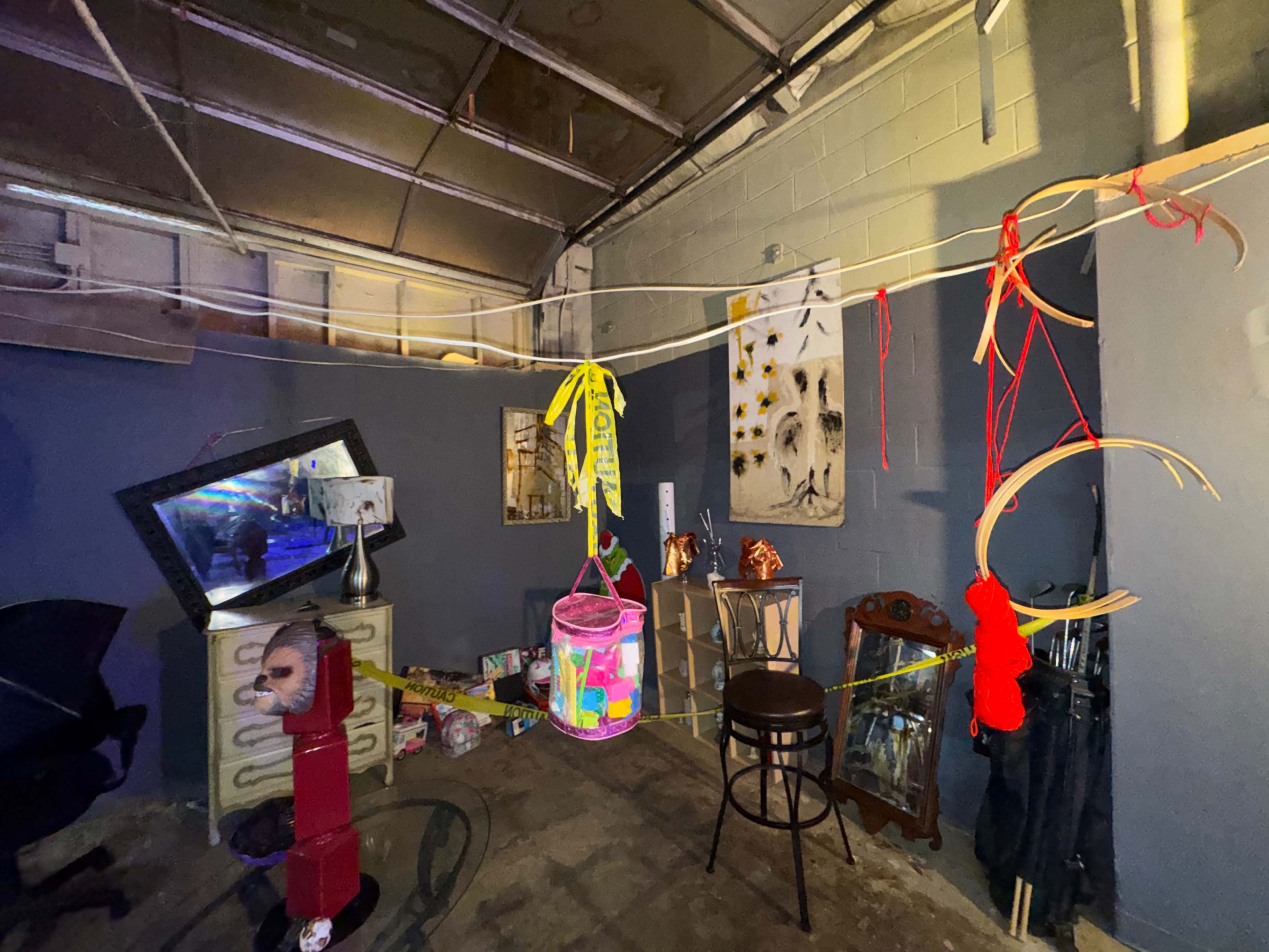 The image shows a dimly lit room with various objects hanging from ropes, including a colorful container, and a mirror reflecting light in the background.