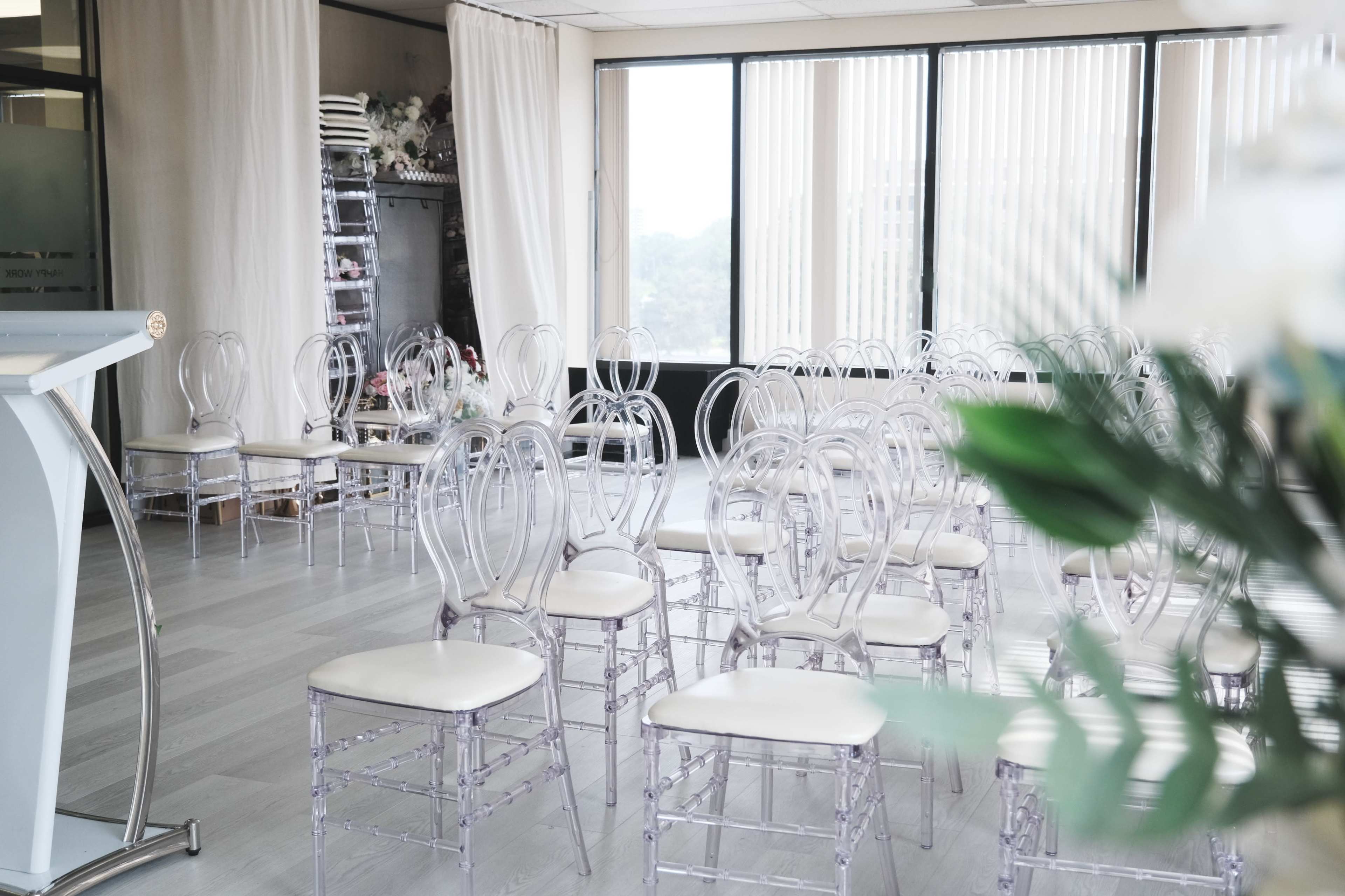 The image shows a setup for an event with clear acrylic chairs arranged in rows inside a room featuring large windows with natural light.