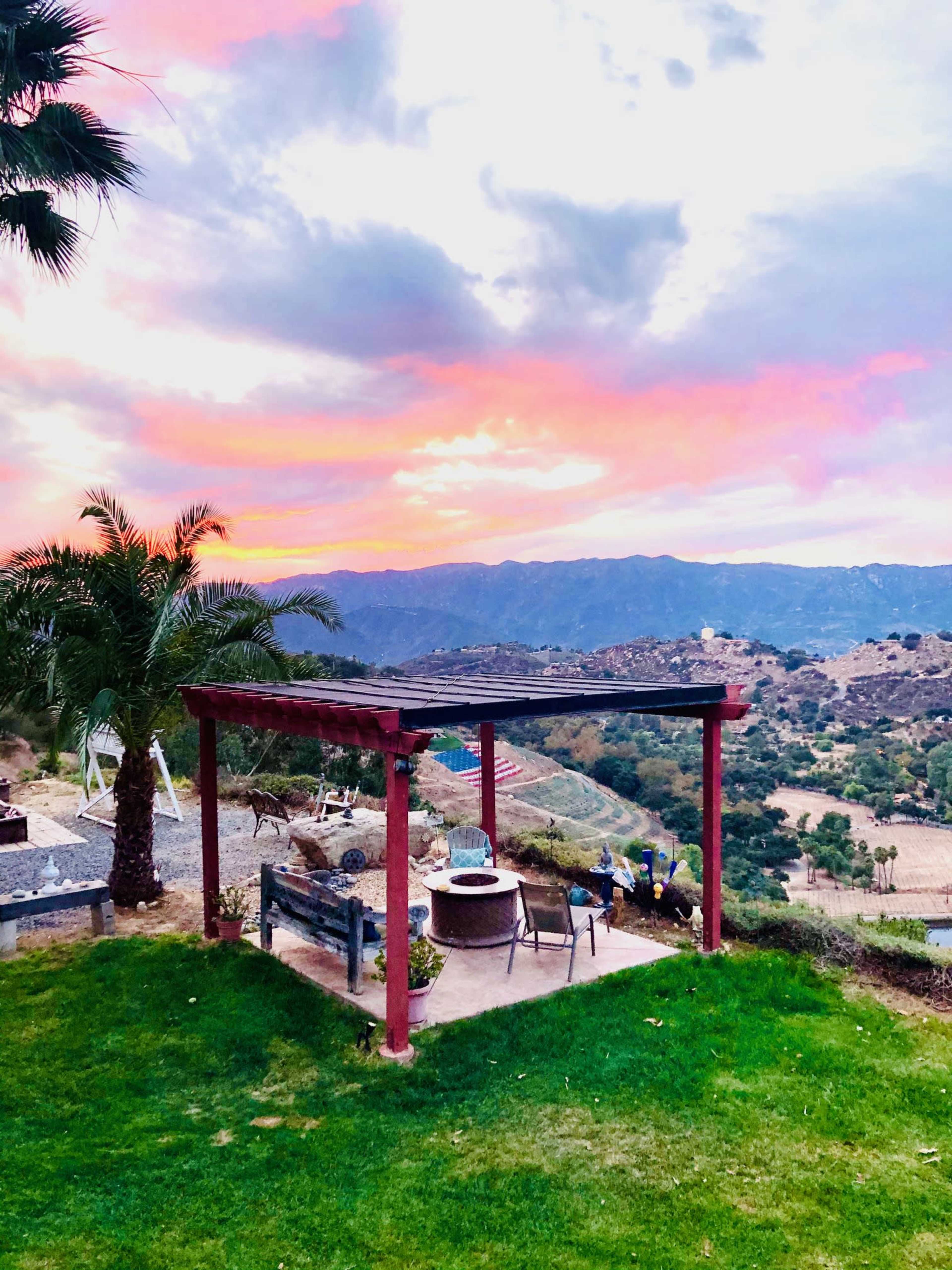 A shaded seating area with a table and chairs is set against a backdrop of mountains and a colorful sunset sky.