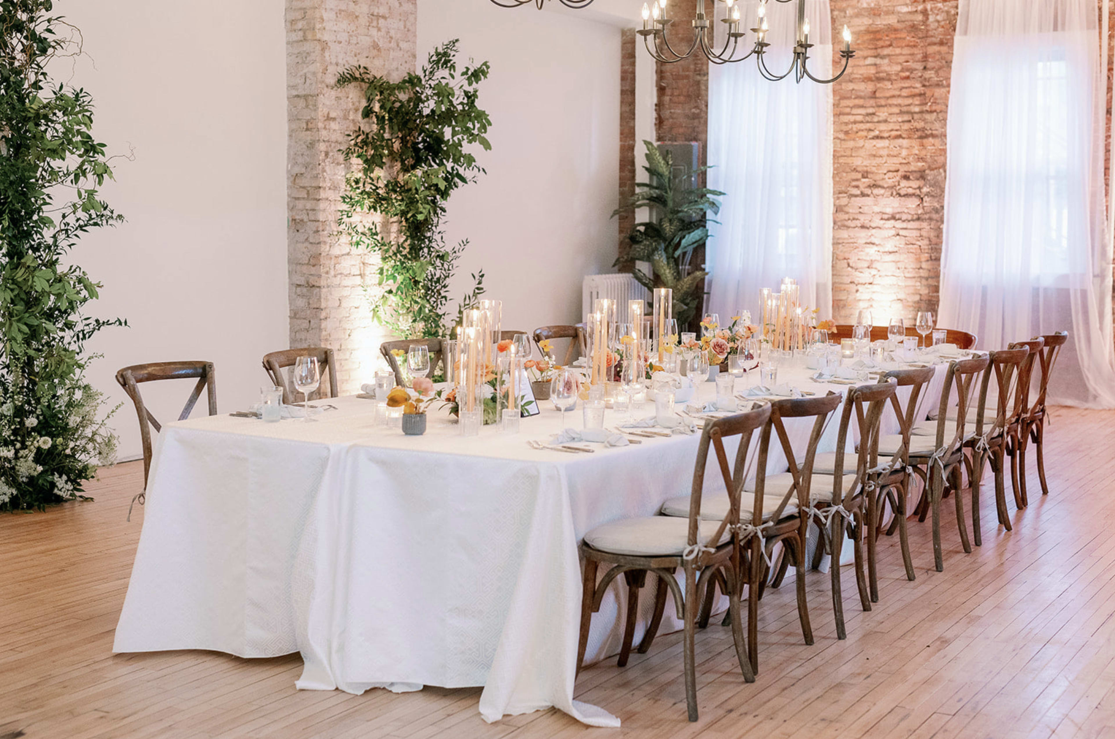 A long dining table is set with elegant tableware and surrounded by wooden chairs in a softly lit room with exposed brick walls.